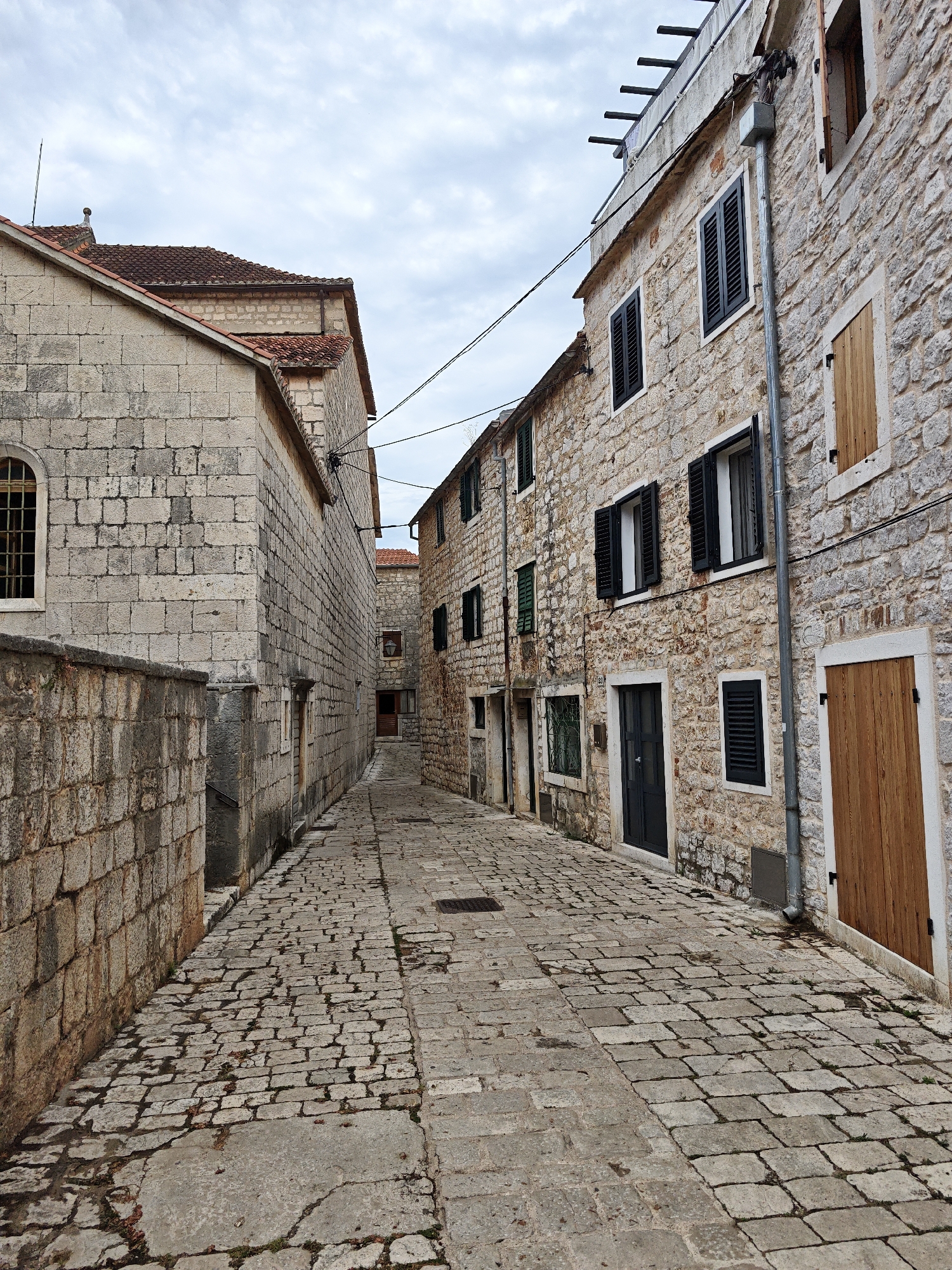 The image depicts a narrow cobblestone alleyway in Grad Stari Grad, Croatia, flanked by old stone buildings. The architecture suggests a historical European setting. On the left, a stone wall separates the alley from a building with a red-tiled roof and a large arched window with visible panes. To the right, a taller stone building features several windows with dark shutters, some open and some closed. A prominent wooden door is visible on the rightmost building. A gray downspout runs vertically along the right building's facade. The cobblestone pavement is uneven and shows signs of wear, with some small leaves and debris scattered across it. The sky is overcast with clouds. There are no people visible in the scene, and no apparent actions or interactions are taking place. The environment is characterized by its age, the stone construction, and the narrow, winding layout typical of old European towns.