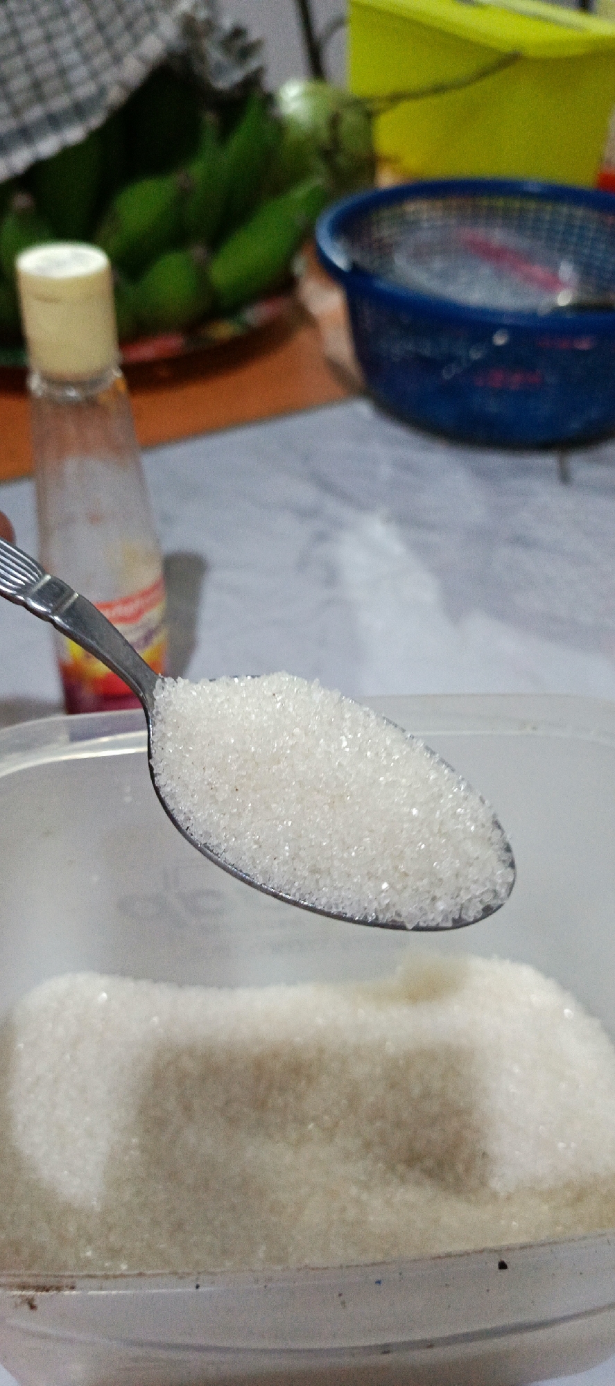 A spoon filled with granulated sugar is held over a clear plastic container also filled with sugar. In the background, out of focus, is a bottle with a yellow cap, a bundle of green bananas, and a blue basket. The setting appears to be a kitchen or food preparation area in Pashiebeutong, Indonesia.