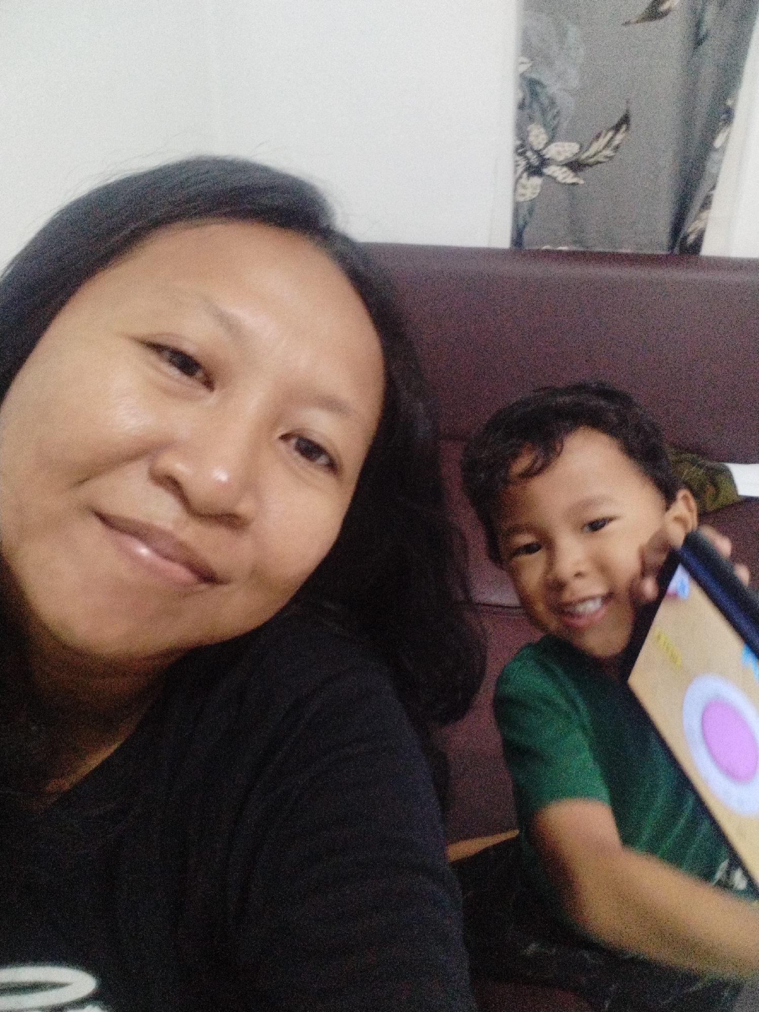 The image captures a selfie taken in what appears to be an indoor setting, possibly a living room, with a woman and a young boy as the primary subjects. The woman is positioned on the left side of the frame, her face filling a significant portion of the left half of the image. She has dark hair, dark eyes, and is looking towards the camera with a slight smile. She is wearing a black t-shirt. To the right of the woman, a young boy is visible. He is smiling and holding up a tablet, which is angled towards the camera. The boy is wearing a green t-shirt. Behind the subjects, a portion of a dark-colored couch is visible, as is a curtain with a floral pattern in shades of gray and white. The lighting appears to be dim, and the image has a slightly grainy texture. The specific location in Karangsentosa, Indonesia, cannot be determined from the visual information alone.