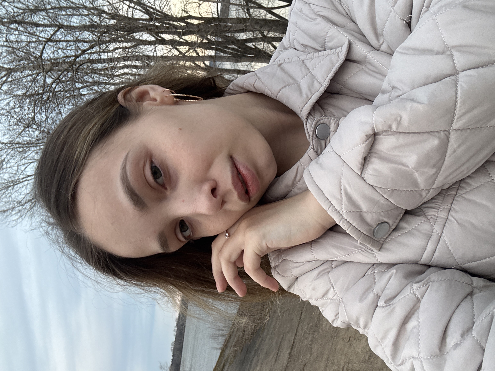 The image presents a close-up, slightly tilted portrait of a young woman, likely a selfie, taken outdoors in a natural setting, possibly in Chaikovsky, Russia. The woman, with light brown hair and a fair complexion, looks directly at the camera with a calm, neutral expression. She is wearing a light-colored, possibly beige or light grey, quilted jacket, which covers her arms and shoulders. Her right hand is gently placed under her chin, resting on her folded arm. A small, delicate ring with a clear stone adorns her right ring finger, and a thin gold hoop earring is visible in her right ear.
The background suggests a cooler season, with intricate patterns of bare tree branches silhouetted against a softly lit sky. Below the trees, a wide body of water, consistent with a river like the Kama in Chaikovsky, stretches horizontally, bordered by a natural, earthy bank in the middle distance. The lighting is soft and diffused, indicating daytime, perhaps early morning or late afternoon, without harsh shadows. The overall scene conveys a quiet, contemplative mood in an outdoor environment. No visible text is present.