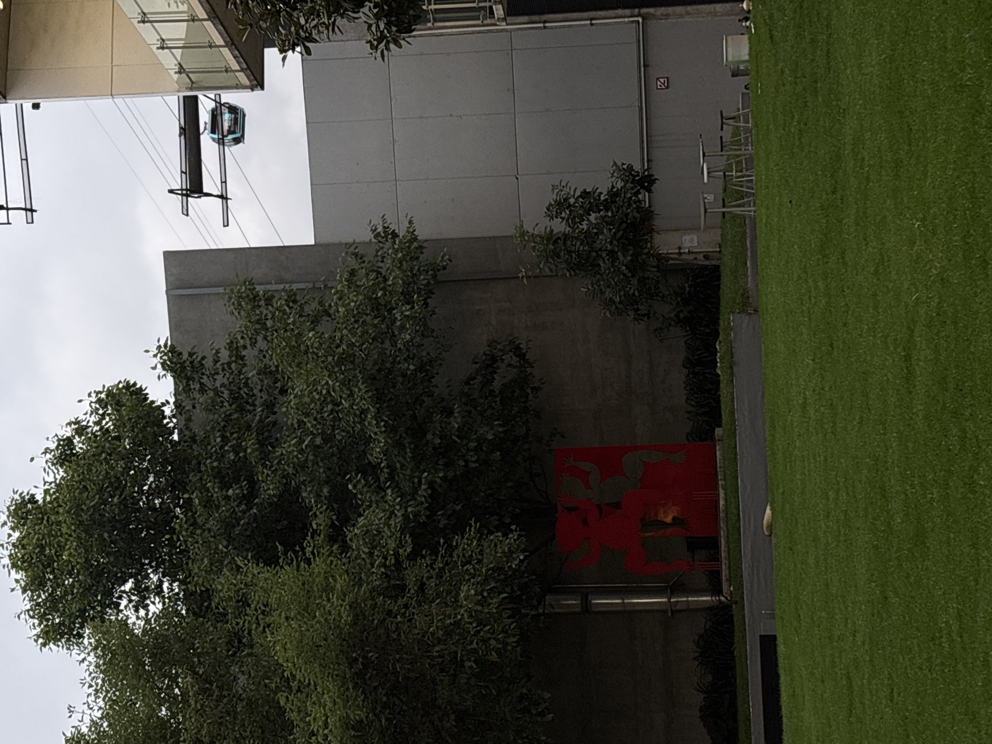 The image shows a cityscape in Mexico City, Mexico. A cable car is visible in the upper left, supported by overhead cables and pylons. Adjacent to the cable car is a building with a gray facade. In the lower center, a lush green tree partially obscures a red wall or structure featuring an artistic design depicting human-like figures. To the right, a manicured lawn extends into the distance. Outdoor seating (tables and chairs) is placed near the building.