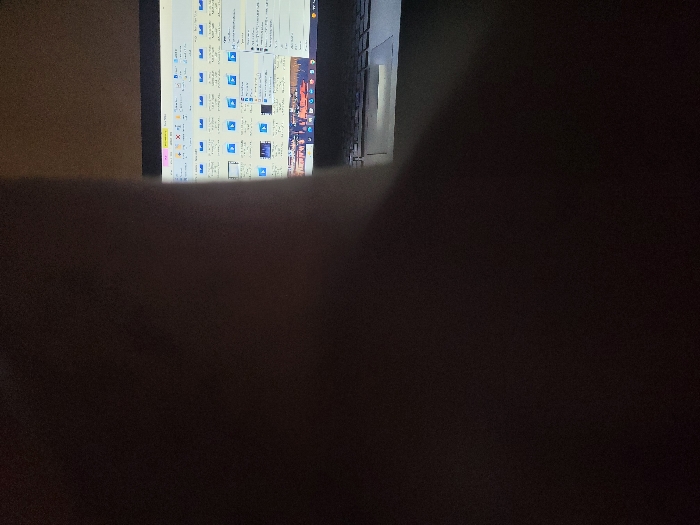 The scene, stated to be located in Kubare, Nigeria, depicts a dimly lit interior environment. The right portion of the image displays a partially visible laptop computer, with its screen illuminated. The screen shows a graphical user interface, likely a file explorer, featuring rows of light blue icons and small thumbnail previews arranged in a grid. Below these, a horizontal section of the screen displays what appears to be a still frame or image, possibly depicting a night scene with multiple artificial light sources, resembling a cityscape or a crowded event. A segment of the laptop's keyboard is visible below the screen. The left and central portions of the image are dominated by a large, amorphous dark brown to black mass, obscuring a significant portion of the foreground and left side of the laptop. No individuals are clearly identifiable, and no direct actions or interactions beyond the display on the laptop screen are discernible.