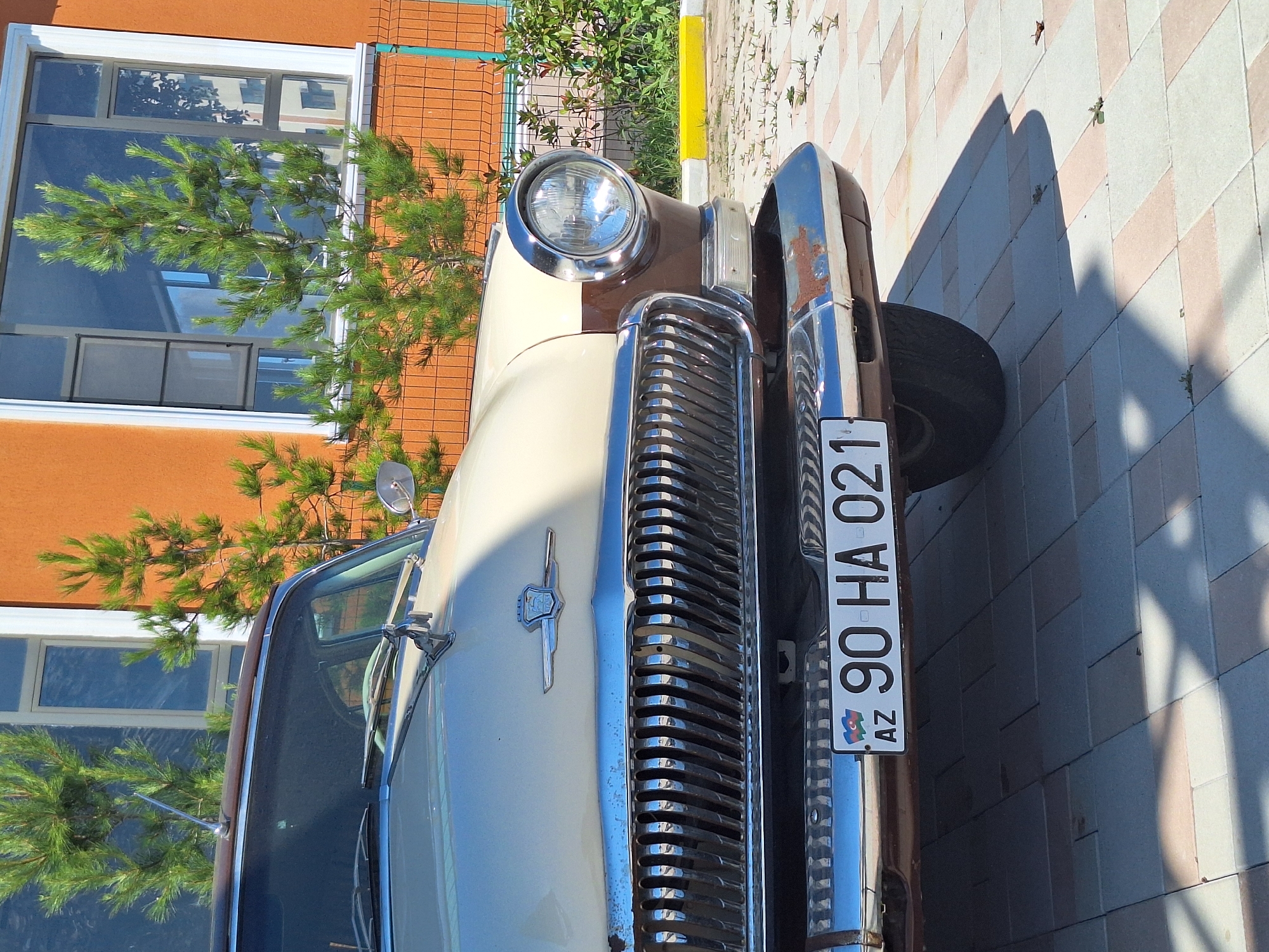 This is a close-up, eye-level, outdoor, medium shot of the front of a vintage cream and brown car. The car's prominent chrome grille dominates the lower half of the image, with vertical slats reflecting light. A license plate reading "AZ 90 HA 021" is mounted on the lower bumper. The car has a large, round headlight with a chrome bezel on the right side. The background features an orange building with a large window, framed by white trim. Several small evergreen trees are planted in front of the building, with a metal fence visible behind them. The ground is paved with light-colored interlocking bricks, and shadows from the car and trees are cast across the surface. The scene appears to be a sunny day, possibly in the city of Khudat, Azerbaijan, given the license plate format.