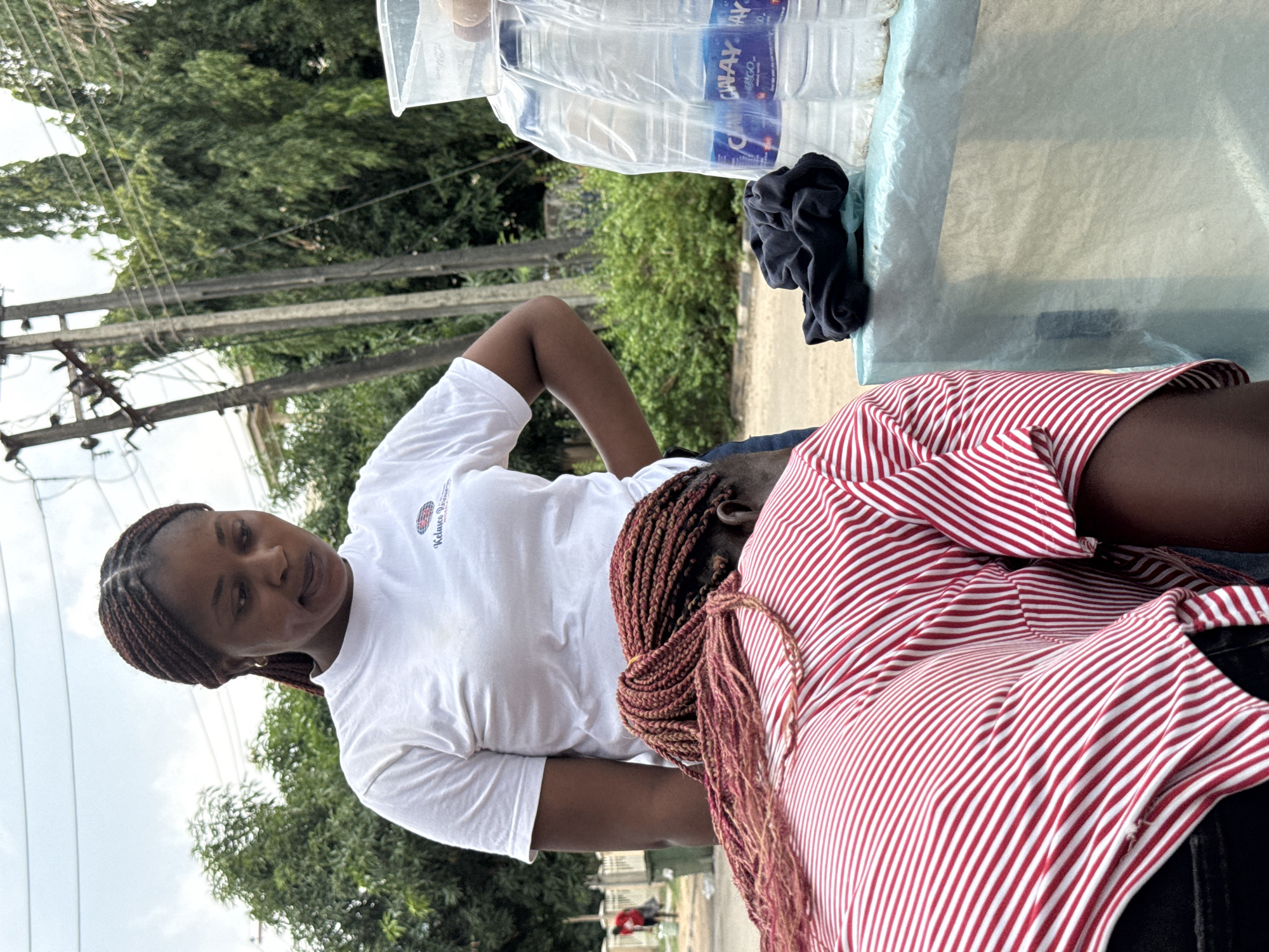 This outdoor, daytime image captures two women interacting in what appears to be an urban or semi-urban setting, possibly in Abuja, Nigeria. The bright lighting suggests it is mid-day with clear weather.
On the left, a woman with dark braided hair stands, wearing a white short-sleeved t-shirt. Visible text on her shirt reads "Kalsuce Pro...". She has a pleasant expression, looking towards the woman on the right, suggesting she is engaging in conversation.
On the right, another woman is seated or leaning, with her back slightly turned towards the viewer, facing the standing woman. She is wearing a red and white striped top, and her light brown braided hair is prominent, extending down her back. Her face is not clearly visible.
Behind the women, on a flat surface (possibly a table or stall), several plastic bottles of water are visible. The brand name "CHIWAY" and "SACO" are discernible on the bottle labels. These bottles are wrapped in plastic packaging. A dark, crumpled piece of fabric is also on this surface.
The background features lush green trees, indicating a natural element within the urban landscape. Utility poles and wires crisscross the upper part of the background, further confirming an outdoor, developed area. A glimpse of the ground or a road is visible in the lower part of the frame. The scene suggests a casual interaction, perhaps during an outdoor event or at a refreshment point.