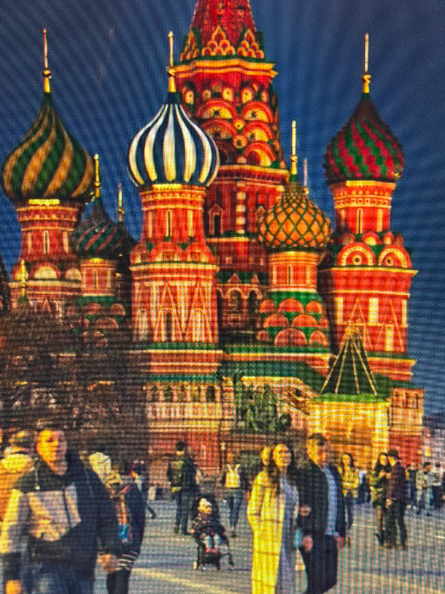 The media file shows a nighttime scene featuring Saint Basil's Cathedral in Moscow, Russia. The cathedral's iconic onion domes are illuminated, with vibrant colors of red, green, and gold against a dark blue sky. In the foreground, a crowd of people are walking and interacting. The people are mostly blurred due to motion and low light conditions, but some individuals are more discernible. A man in a dark jacket and jeans is prominent, walking towards the viewer. A woman in a yellow coat is walking alongside him, with her arm linked through his. Other figures are scattered throughout the scene, including a child in a stroller. The overall atmosphere appears to be one of a public gathering or tourist activity at dusk or after dark. There is no visible text in the media file.