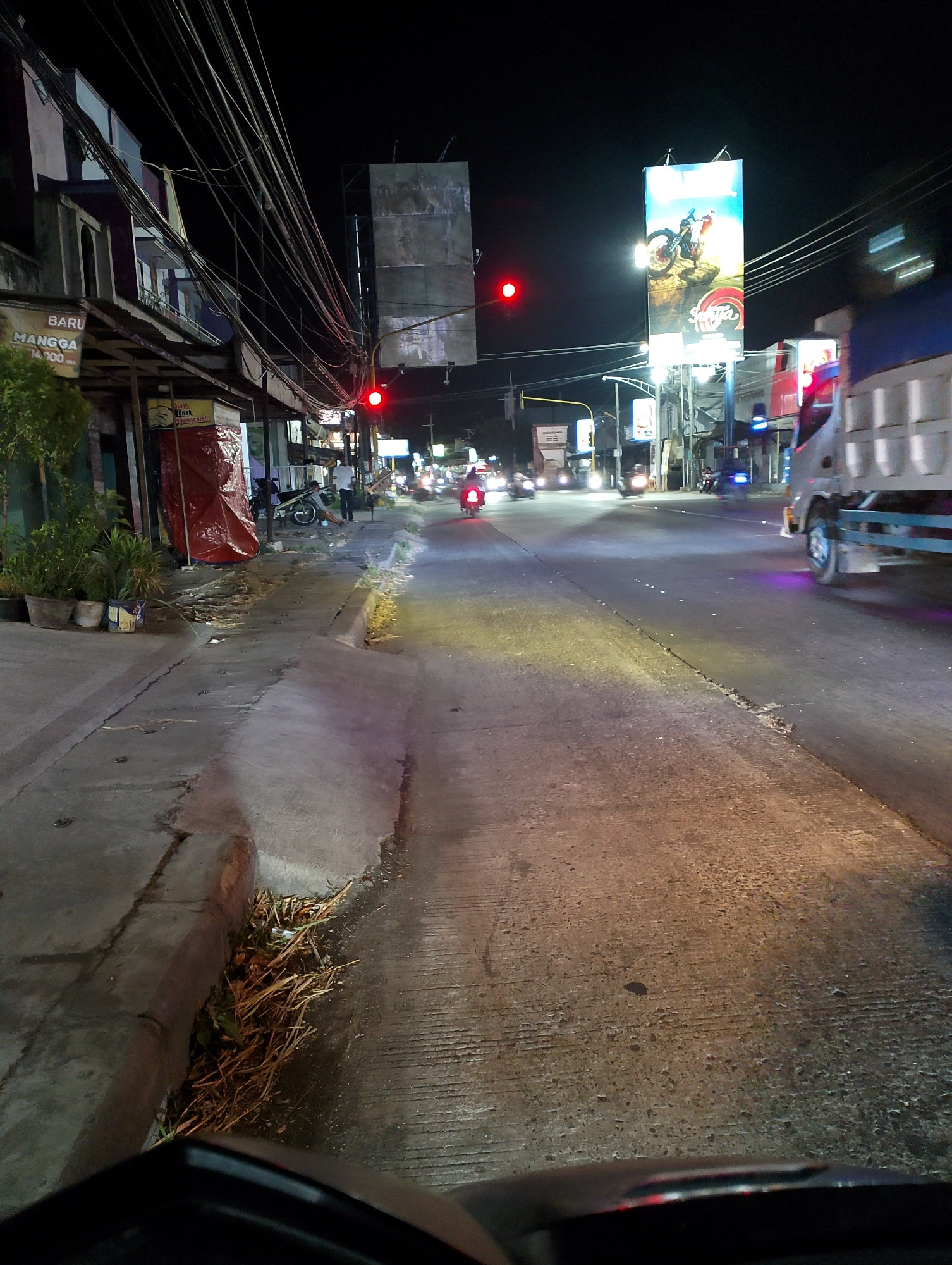 The image depicts a street scene at night in Gringging, Indonesia. A red traffic light is visible on the left side of the road. Several buildings line the left side of the street, with awnings and signs. One sign reads "BARU MANGGA" and another, partially obscured, appears to advertise a business. A red tarp is draped over something on the left sidewalk. A motorcycle is parked on the sidewalk near the buildings.

The road is paved and appears wet in places, reflecting the streetlights and vehicle lights. A large billboard advertising a motorcycle is visible on the right side of the road. A white truck is moving away from the camera on the right side of the road. Other vehicles, including motorcycles, are visible further down the street. The sky is dark, indicating it is nighttime. The foreground shows a portion of a vehicle, possibly a motorcycle, from the perspective of the rider. The overall scene conveys a typical urban street at night with moderate traffic.