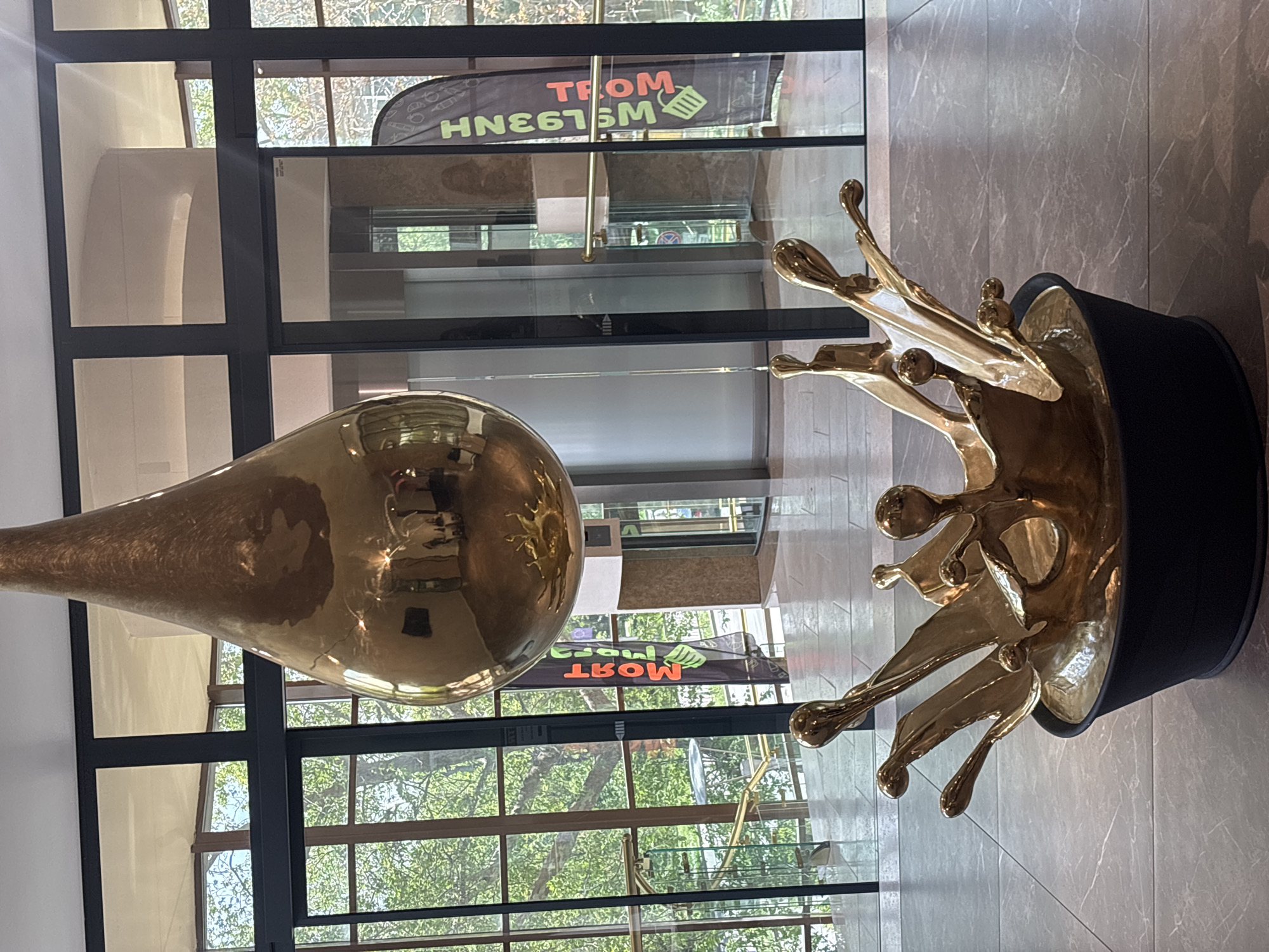 The image captures an indoor scene, likely within a commercial or public building in Varna, Bulgaria, as suggested by the reversed text "Most Varna" visible on a sign outside the windows. Two modern art pieces are the primary focus. On the left, there's a large, elongated, pear-shaped, reflective, golden object suspended from the ceiling. To the right, a golden sculpture resembling a crown with fluid, splash-like extensions sits on a black cylindrical base. The objects are positioned against a backdrop of glass windows and black framing, providing views of greenery and the exterior. The interior features a light-colored tiled floor and neutral-toned walls. The reflective surfaces of the golden objects capture elements of the surrounding environment, including architectural details and people's silhouettes.