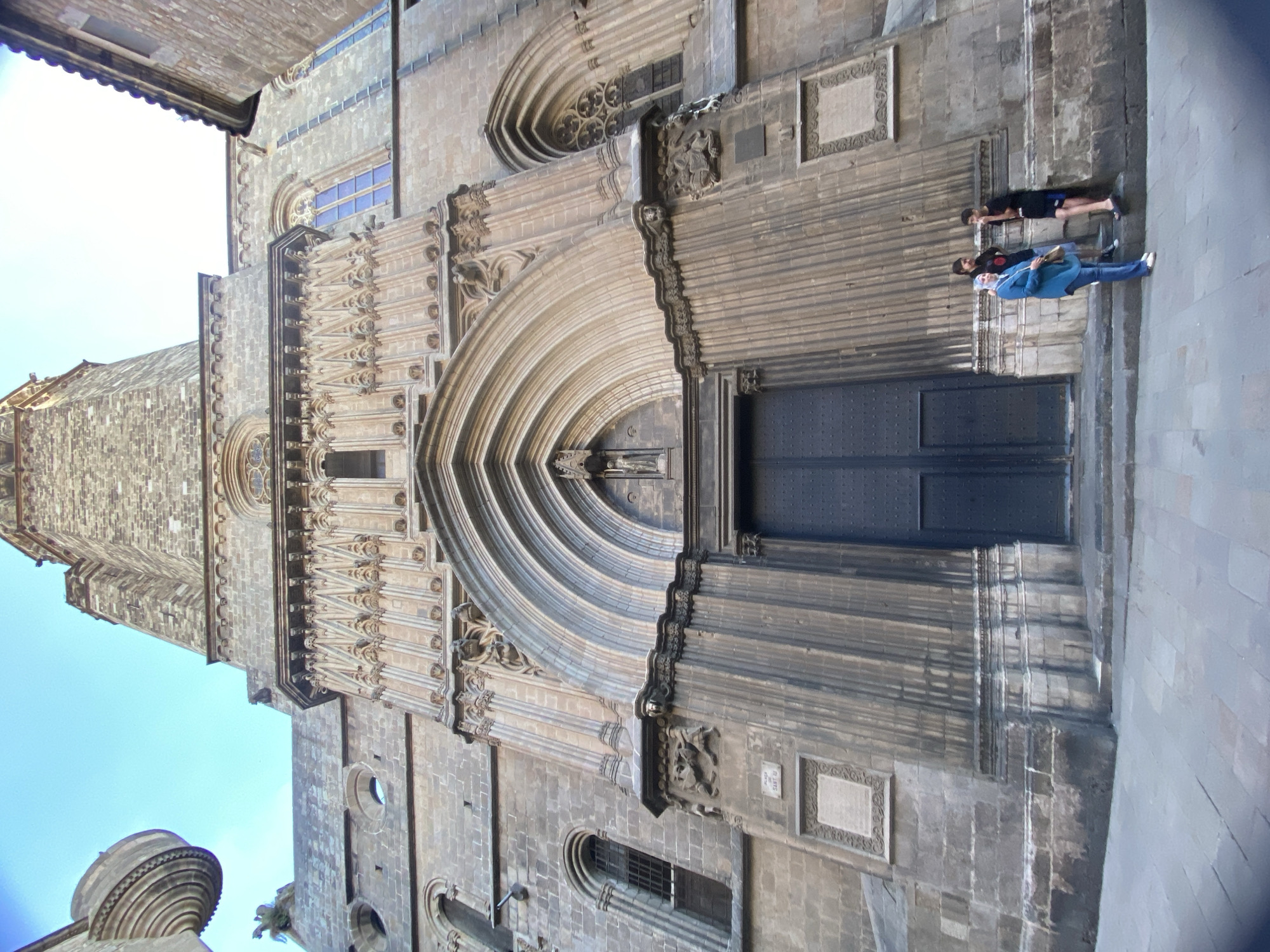 A low-angle, wide shot captures the imposing facade of the Barcelona Cathedral in Barcelona, Spain. The gothic architecture is characterized by intricate stonework, pointed arches, and ornate carvings. Three individuals are present, standing on the steps leading to the cathedral's large, dark wooden doors. One person, wearing a blue headscarf and tunic, stands to the left, facing the camera. Two others, appearing to be young males, are positioned to the right. One is leaning against the wall, smoking, while the other stands in conversation. The sky above is clear and blue. The cobblestone pavement in the foreground suggests a public square or plaza.