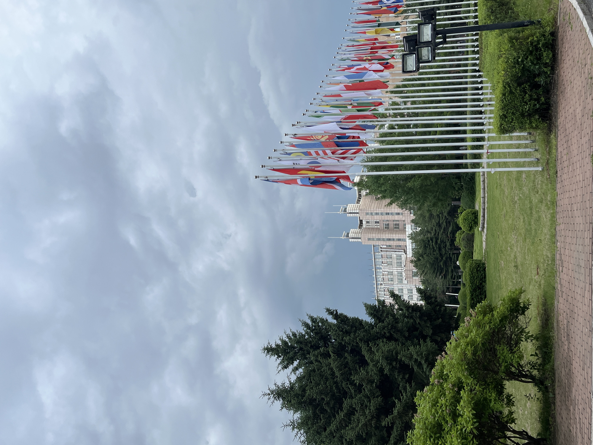 The image depicts a cloudy day in Changchun City, China, likely at the United Nations Building, a landmark known for its extensive flag display. In the foreground, a red brick pathway leads to a manicured lawn with lush green bushes and trees. Behind the greenery, a row of white flagpoles stands tall, displaying a variety of national flags fluttering in the wind. These flags are arranged in a semi-circular fashion, creating a dynamic visual effect. In the background, a large, modern building with a light-colored facade and multiple windows dominates the skyline. The sky is overcast with heavy, dark clouds, suggesting a possibility of rain. The overall scene conveys a sense of international gathering and architectural grandeur, characteristic of a significant government or diplomatic building.