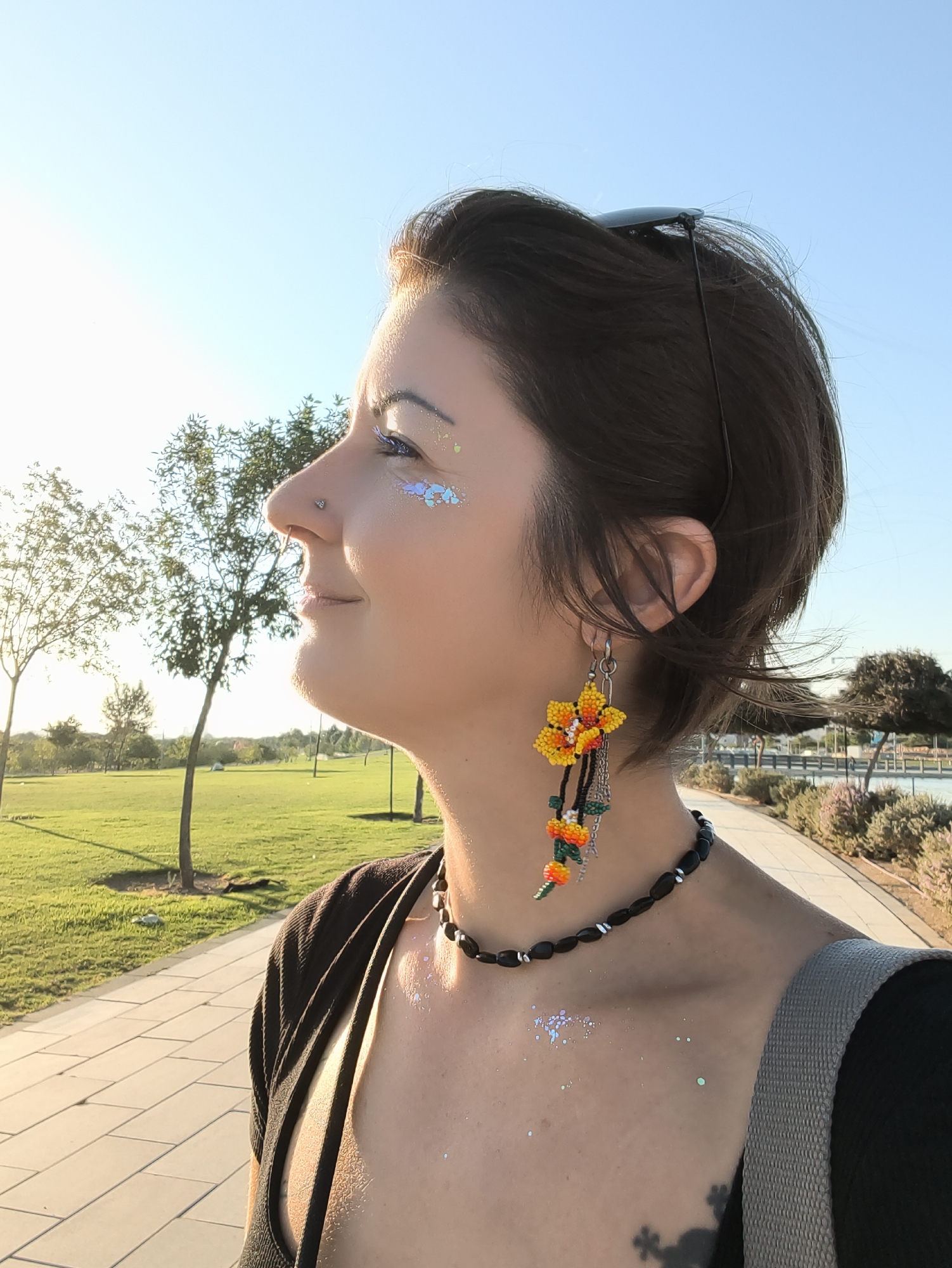 A medium shot shows a woman in profile, looking to the left, with a bright, clear sky behind her and a grassy park with trees and a paved path in the background. The sun is low in the sky, suggesting it is late afternoon or early evening, casting a warm glow on the scene. The woman has short, dark hair and is wearing large, colorful beaded earrings shaped like flowers and a beaded necklace. She has glitter on her face and chest. She appears to be outdoors, possibly in a park or recreational area, given the presence of trees, grass, and a paved walkway. The setting seems peaceful and relaxed. The woman's gaze is directed towards something out of frame, and her expression is neutral or contemplative. There is no visible text in the image.
