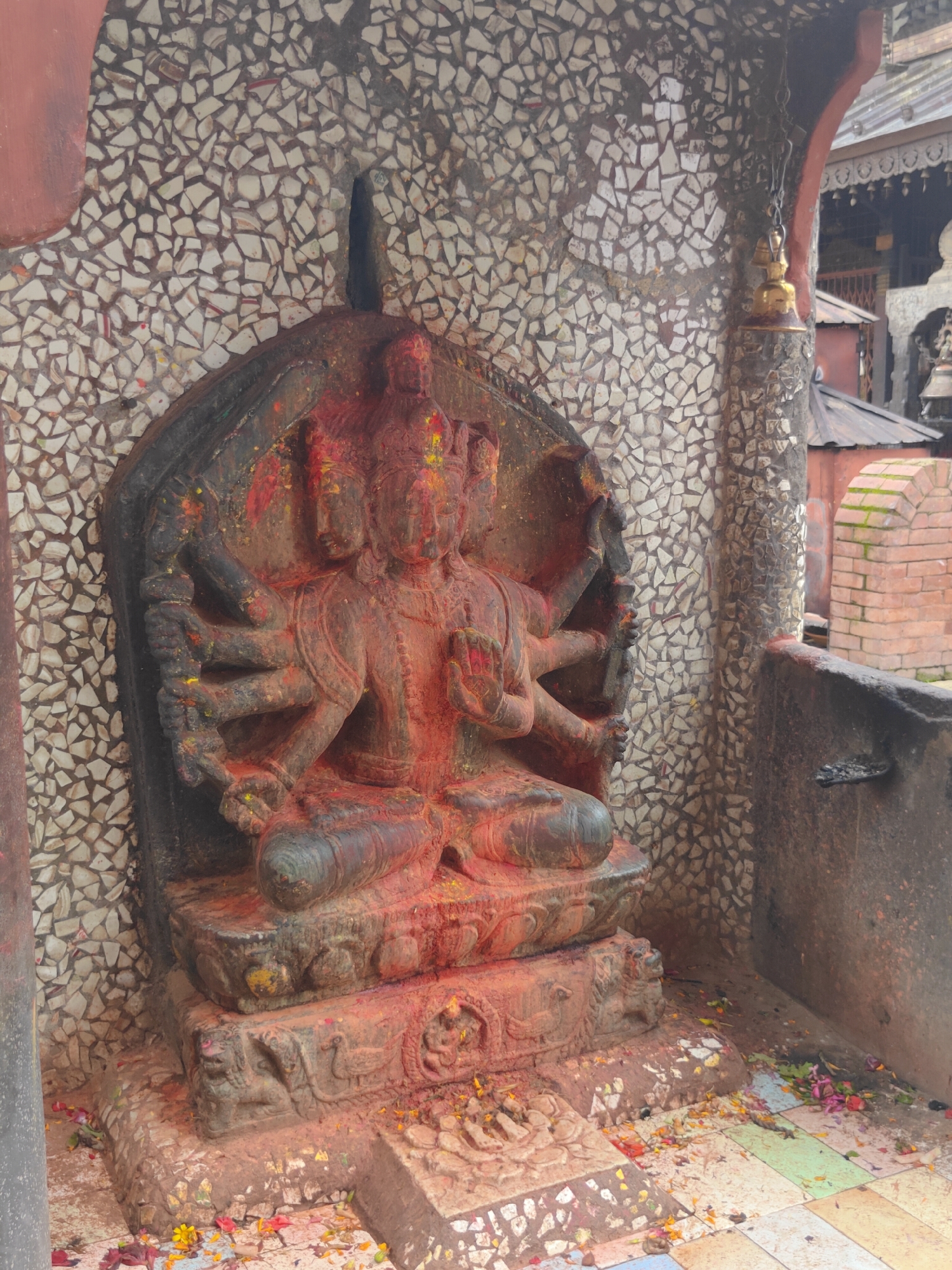The image shows a stone statue of a multi-armed deity, likely Hindu, at the Pashupatinath Temple in Kirtipur, Nepal. The statue is adorned with red powder, possibly vermilion or sindoor, and appears to be a central religious artifact. It is seated on a decorative stone pedestal carved with animal figures, and the surrounding area is littered with fallen flower petals. The statue is set against a textured wall made of small, irregular white and grey tiles. In the background, parts of the temple complex are visible, including brick structures and hanging bells. The overall atmosphere suggests a place of worship, with evidence of offerings and devotional practices.
