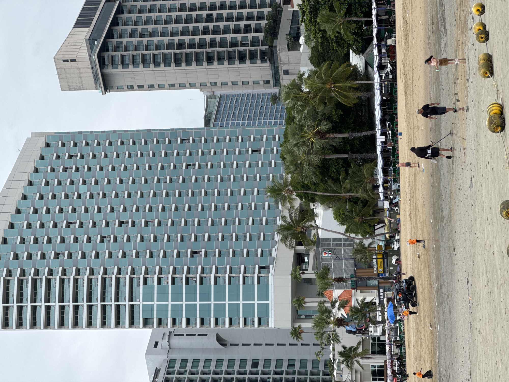 The image depicts a wide, sandy beach in Pattaya City, Thailand, under an overcast sky. In the foreground, the wet sand is interspersed with shallow pools of water, and a rope with several yellow buoys stretches across the lower portion of the frame.
Several people are present on the beach. In the mid-ground, two individuals, possibly beach cleaners wearing orange vests, are working. One is pushing a utility vehicle that has an orange umbrella attached. Another person in an orange vest is seen using a broom to sweep the sand. Further back, a group of people are gathered near beach umbrellas and chairs.
In the background, a row of tall, modern buildings dominates the skyline. The most prominent building is a large, light blue and gray skyscraper with a repetitive pattern of balconies and windows. To its right is another tall, light-colored building with a similar architectural style. Further in the distance, more buildings are visible. Palm trees are scattered throughout the scene, adding to the tropical setting. The overall impression is a bustling urban beach with a mix of leisure activities and city infrastructure.