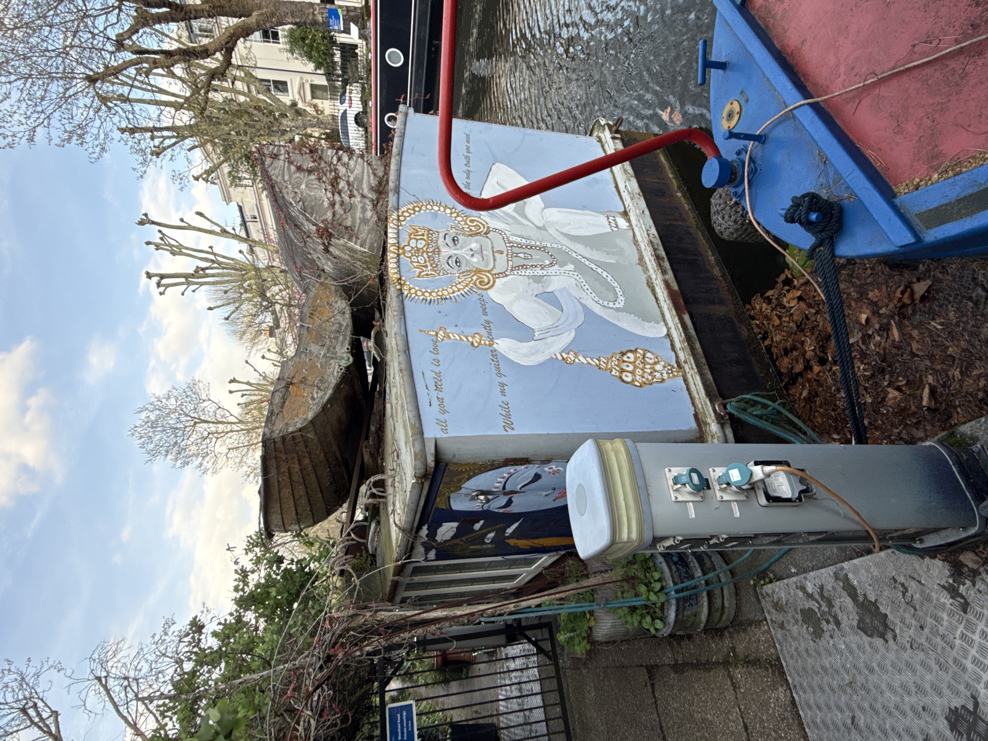 Here is a detailed description of the image:
**Scene Overview:**
The image appears to be taken outdoors alongside a canal in London, United Kingdom, likely in a residential area. The scene features a collection of objects and structures related to a houseboat or canal boat docked along the canal's edge.
**Key Elements:**
* **Canal Boat/Houseboat:** Several sections of a boat are visible. One section is brightly painted with a blue background and an image of a figure (likely an angel or deity) with ornate detailing. There is also text near the figure's head and body, including the phrase "all you need is love."
* **Boat Exterior:** Adjacent to the painted section is what appears to be the weathered, curved roof or hull of another boat section. The roof appears to be constructed from aging wood or a similar material.
* **Mooring Point/Electric Supply:** A gray rectangular unit with electrical sockets is prominent in the foreground, placed next to the boat. This suggests the boat is docked at a designated spot with electrical hookups.
* **Water:** The canal's water is partially visible in the upper right section of the image.
* **Foreground Elements:** The foreground includes an aluminum plate surface with a diamond pattern and potted plants.
* **Vegetation:** The image captures glimpses of vegetation including bushes and tree branches.
* **Background:** The background reveals portions of buildings, trees, and a cloudy sky.
**People:**
There are no people present in the image.
**Overall Impression:**
The image gives the impression of a lived-in space on a canal boat, combining artistic elements with the more functional aspects of mooring. The canal side setting and diverse elements give a glimpse into an alternative urban lifestyle.
