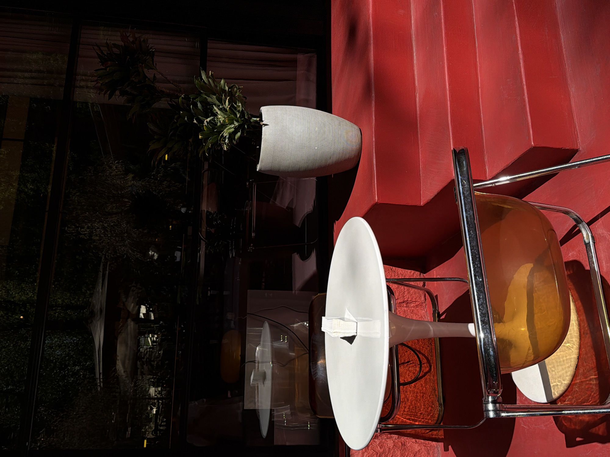 A horizontally-oriented, color photograph depicts a still life scene. A plant, potted in a gray, cylindrical vessel with horizontal ridges, is mounted on a red wall. A chair, constructed with a chrome frame, features a transparent amber seat and a white, oval backrest secured with white tape. The scene is partly reflected in a glass surface, revealing an exterior scene of a windowed building. The red wall has a stepped, geometric texture. While the city is not directly depicted, Yerevan, Armenia is noted as the location. There are no people visible.