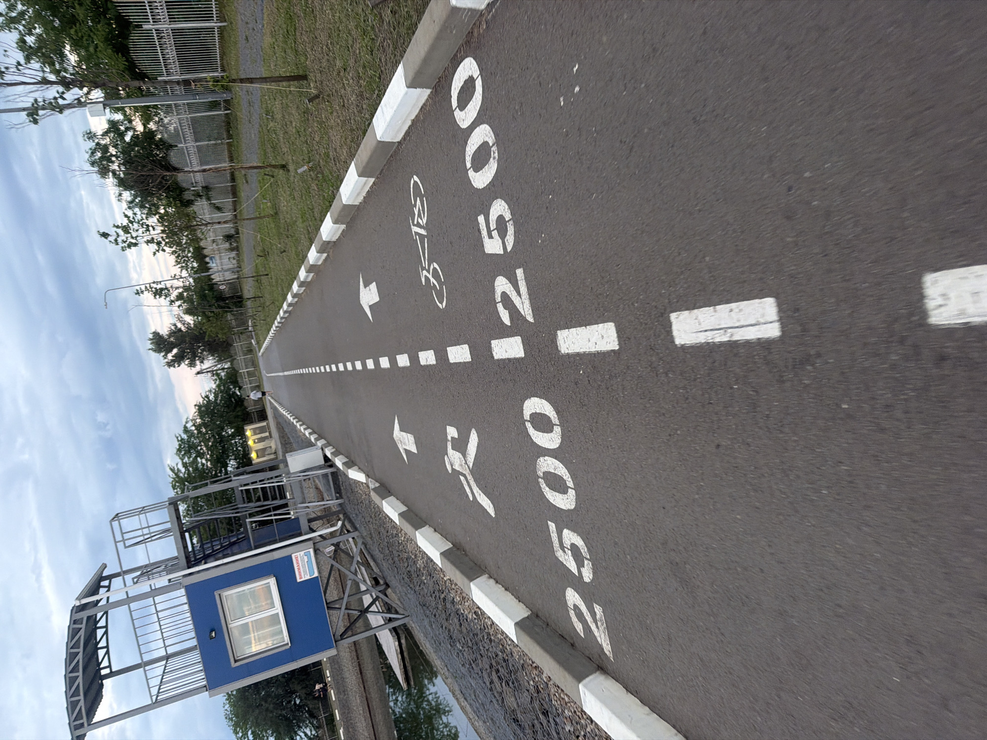 An outdoor asphalt pathway, designated for cycling and running, stretches into the distance under a partially cloudy sky, likely captured in the late afternoon or early evening. The path is marked with clear white paint, including dashed lines, directional arrows, a bicycle symbol, and a running person symbol. Prominently displayed numerical markings "12500" and "2500" are visible on the asphalt, suggesting distance markers, possibly in meters.
The pathway is bordered on one side by a grey and white checkered curb, beyond which lies a strip of green grass. A metal fence runs parallel to the track, separating it from an area with several young trees. In the mid-ground on the left, a tall, multi-level structure made of metal scaffolding supports a blue booth with a window. On the side of this blue booth, a red sign with white text reads "ВНИМАНИЕ!" (Vnimanie!), which means "ATTENTION!" in Russian, possibly indicating a warning or information point for users of the track. The entire scene points to a dedicated recreational or sports facility in Rostov-on-Don, Russia, with clear guidance for participants.