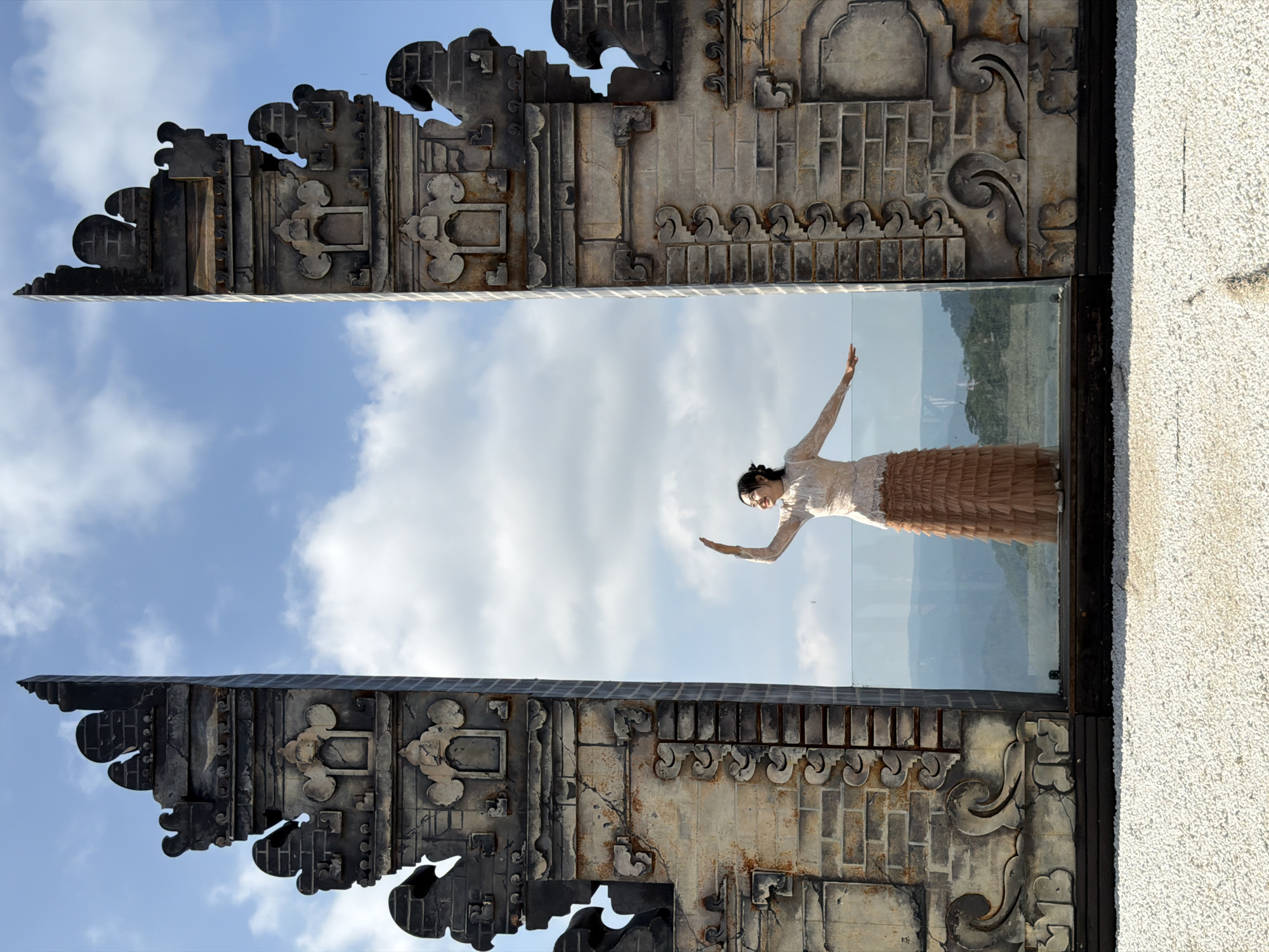 The image shows a woman standing in front of a large, ornate gateway in Da Lat, Vietnam. The gateway is made of stone and features intricate carvings. The woman is wearing a long, flowing dress and has her arms outstretched. She is looking up at the sky. Behind the gateway, there is a panoramic view of the lush, green hills and mountains surrounding Da Lat. The sky is blue with white clouds. The overall impression is one of peace and tranquility.
