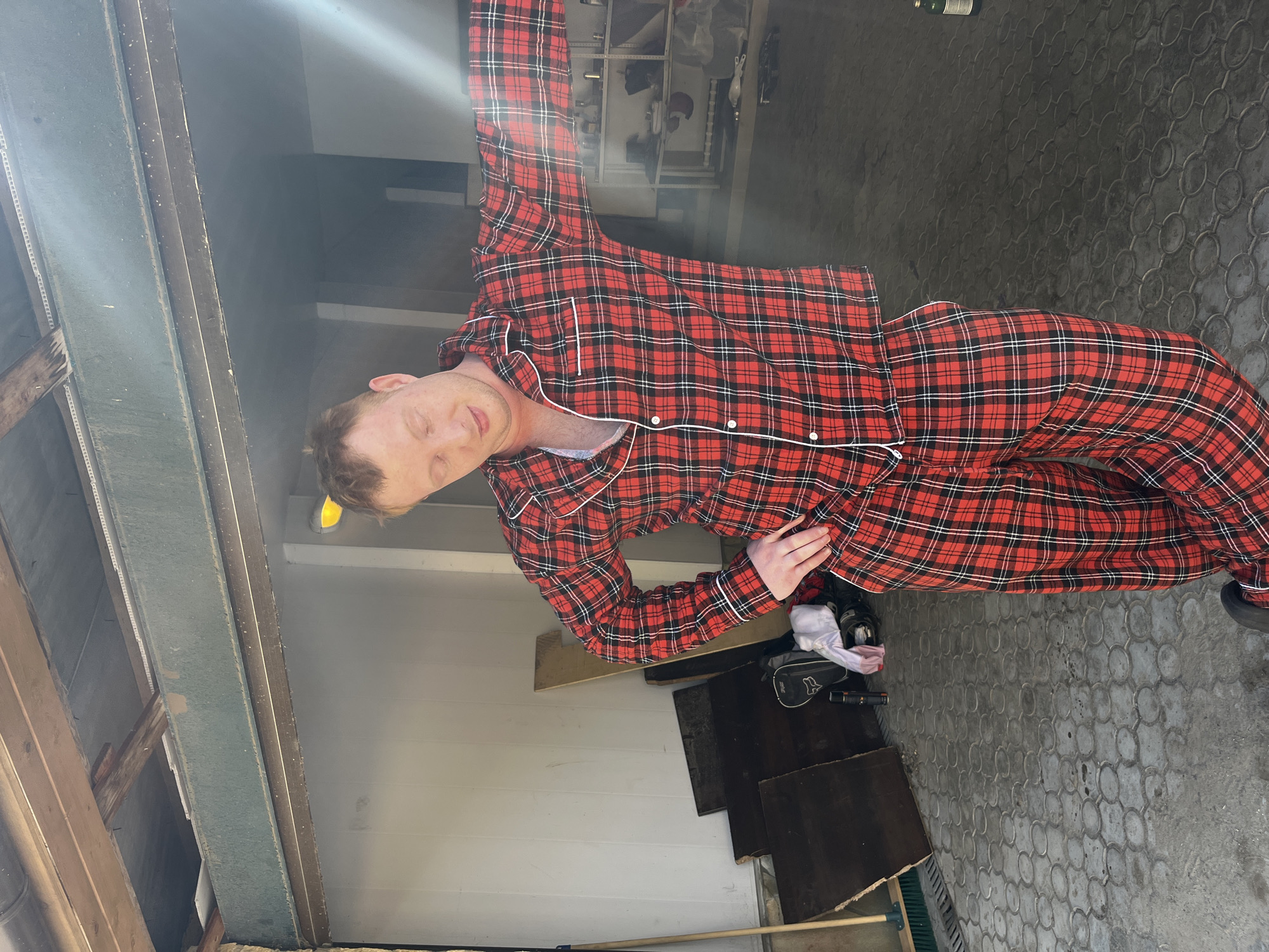 A red-haired person wearing red and black plaid pajamas stands in what appears to be a garage or storage area. The floor is concrete with a circular pattern. To the left, the garage door opening is visible. A white light illuminates the ceiling and upper left corner of the scene. Miscellaneous items, including boards and containers, are scattered on the floor near the center of the image. The person's eyes are closed. The location, Ореховка, Russia, is not discernible from the image.