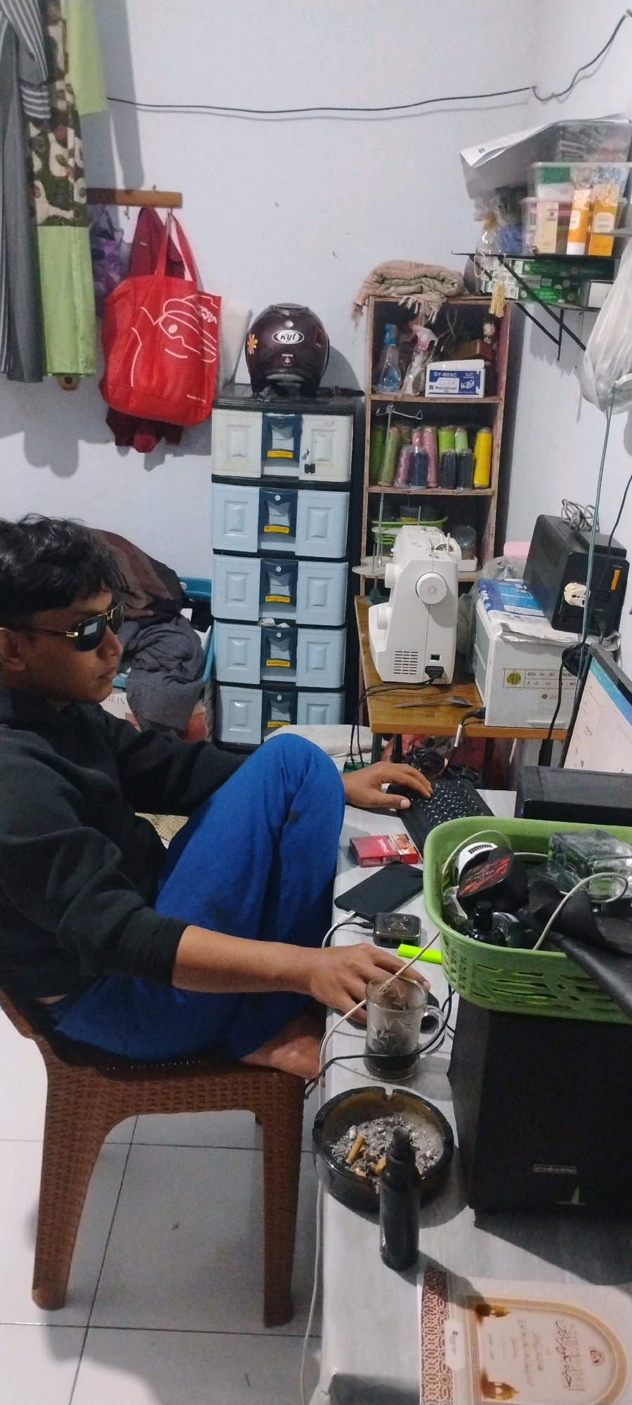 In a room, a young male wearing sunglasses and a black hoodie sits on a woven plastic chair with his legs crossed. He is looking at a computer screen and typing on a black keyboard, with his right hand resting on the mouse. On the table in front of him are a pack of cigarettes, a smartphone, a green plastic basket containing various items including a computer mouse, and a glass mug with a beverage. To his right, there is an ashtray with cigarette butts and a black bottle, and a large black speaker. Behind him, a white sewing machine sits on a wooden desk, next to a stack of boxes and a shelf unit filled with spools of thread and other items. Further to the left, a red shopping bag hangs on a hook next to a helmet placed on top of a chest of drawers. The walls are painted white, and the floor is tiled. The scene appears to be a personal workspace, possibly in a residential setting in Mumbulsari, Indonesia.
