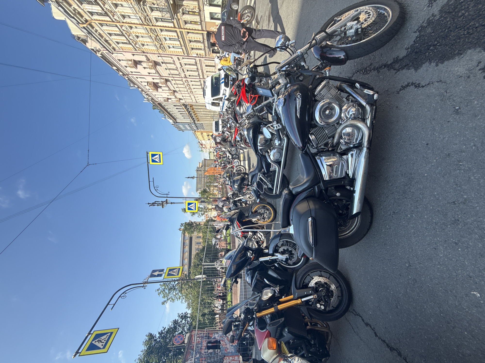 This is a street-level, medium-distance, eye-level shot of a gathering of motorcycles parked on an asphalt street in Saint Petersburg, Russia. The day is sunny with a clear blue sky. In the foreground, several motorcycles are prominently displayed, including a black cruiser with chrome accents and saddlebags, and a red and black sportbike. Further back, a police officer in a black uniform with "МВД РОССИИ" (Ministry of Internal Affairs of Russia) written on his back stands to the right of the frame, facing away from the viewer. Behind the police officer, a white van is parked. A variety of other motorcycles are parked behind the police officer and van. Pedestrian crossing signs with yellow diamond shapes and white pedestrian silhouettes are visible on poles above the street. In the background, ornate buildings with arched windows and balconies line the street, typical of Saint Petersburg architecture. Trees and a metal fence are also visible in the mid-ground. The overall scene suggests an organized event or rally.