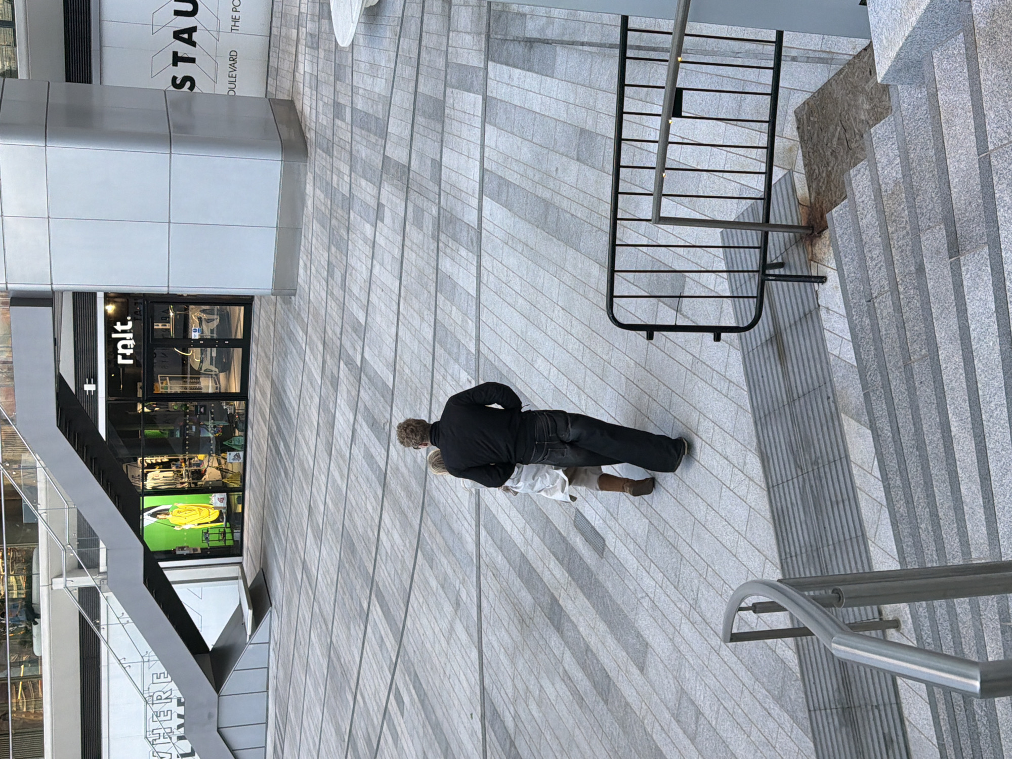 A street-level shot depicts two individuals walking on a textured, paved area in what is identified as the City of London, United Kingdom. A man, dressed in a black jacket and dark trousers, is positioned slightly ahead of a woman. The woman is dressed in a white coat and brown boots. They are walking towards a staircase with metal railings. Adjacent to the paved area, a building with large windows displays the storefront "rlt". Nearby the building is a black metal barricade. The area is primarily composed of gray brick or stone.