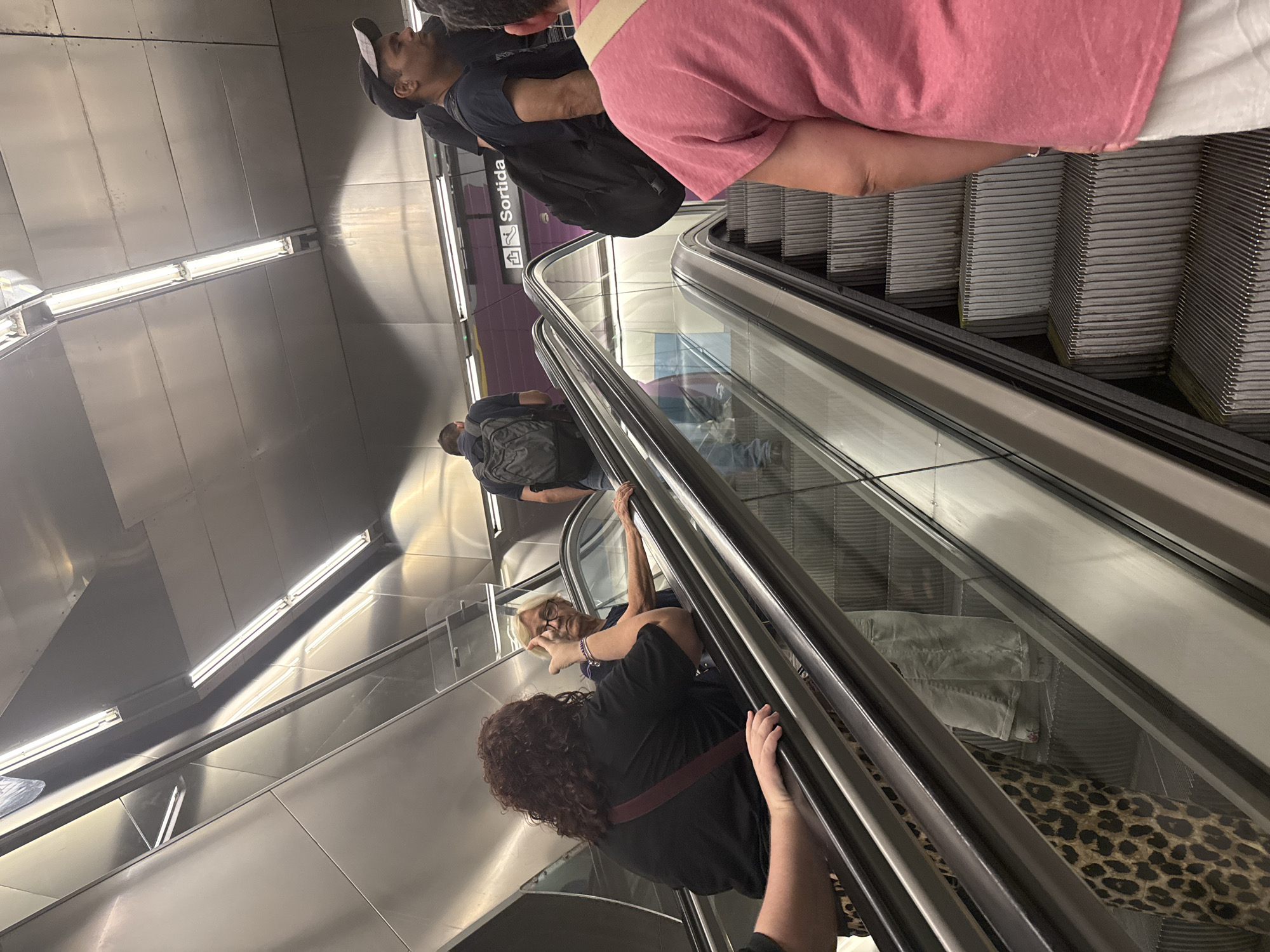 The image shows a group of people on an escalator in what appears to be a metro station. The sign "Sortida" indicates "Exit" in Catalan and Spanish, suggesting the location is in Barcelona, Spain. In the foreground, a person wearing a pink shirt and white pants stands on the escalator. To their left, a woman with curly brown hair and a black shirt is also on the escalator, facing another woman with blonde hair and glasses. Further up the escalator, a man with a backpack is visible, as is another man wearing a black cap and a black t-shirt. The escalator is made of metal with ribbed steps, and the walls are lined with brushed metal panels. Fluorescent lights illuminate the area from above.