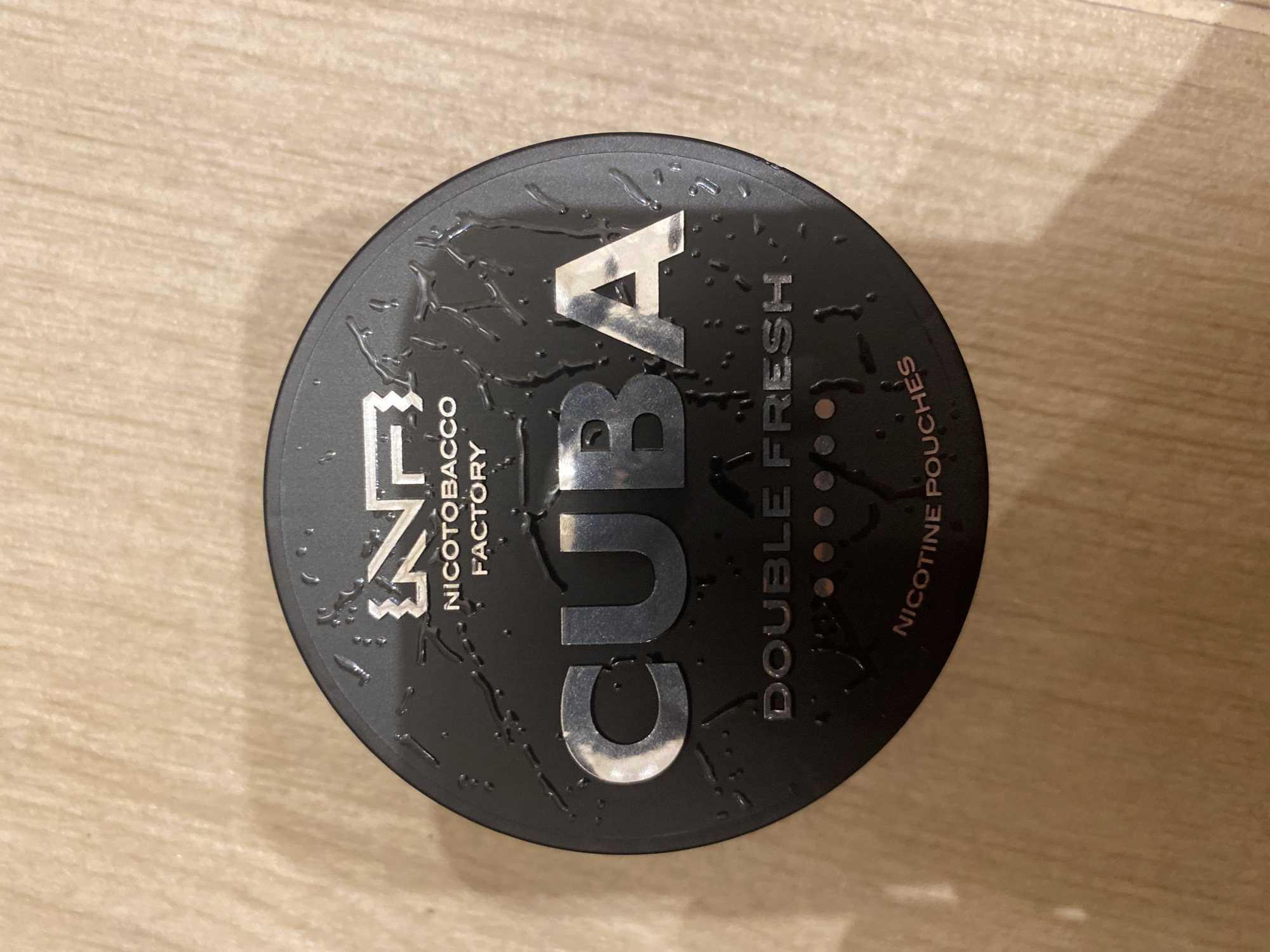 A close-up, overhead shot shows a black, round container labeled "CUBA" in large, silver letters. The container also reads "NICOTOBACO FACTORY," "DOUBLE FRESH," and "NICOTINE POUCHES." Water droplets are visible on the lid. The container rests on a surface with a wood-like grain. There is no evidence to indicate the location as Sofia, Bulgaria.