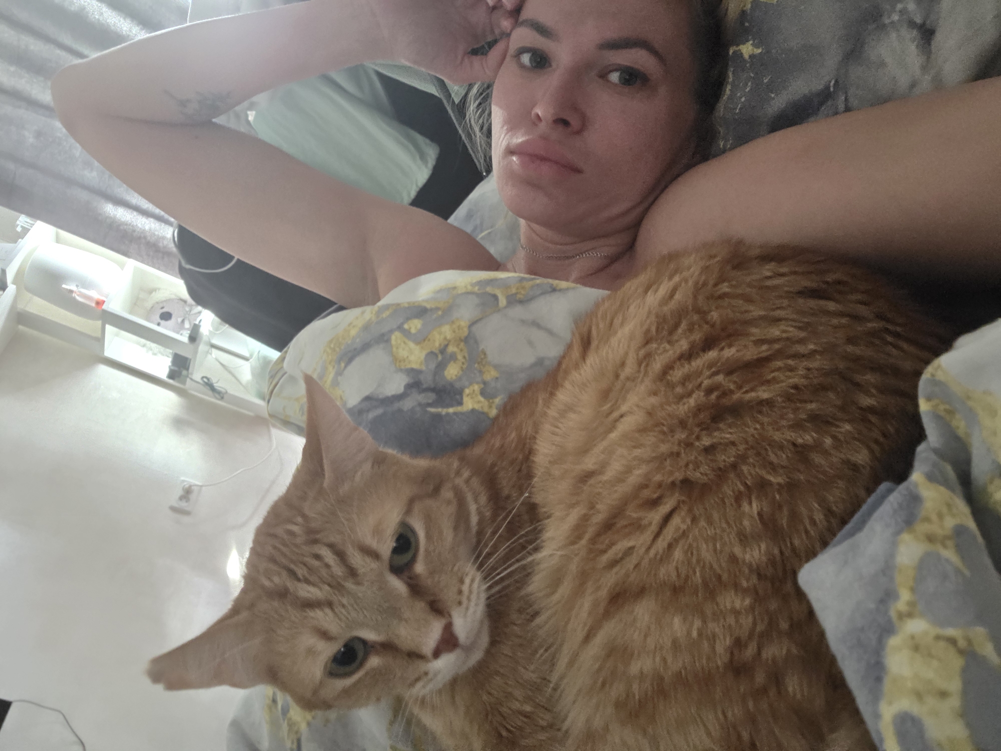 A woman is lying in bed next to a large orange tabby cat. The woman is fair-skinned with blonde hair and is looking directly at the camera with a neutral expression. Her left arm is raised and her hand is resting on her head. The cat is lying close to her, with its head resting on the pillow. The pillow has a grey and gold pattern. The bed has grey and gold sheets. There is a table next to the bed. The photo appears to be taken indoors, and the room is lit by natural light. The city is not discernible from the image.