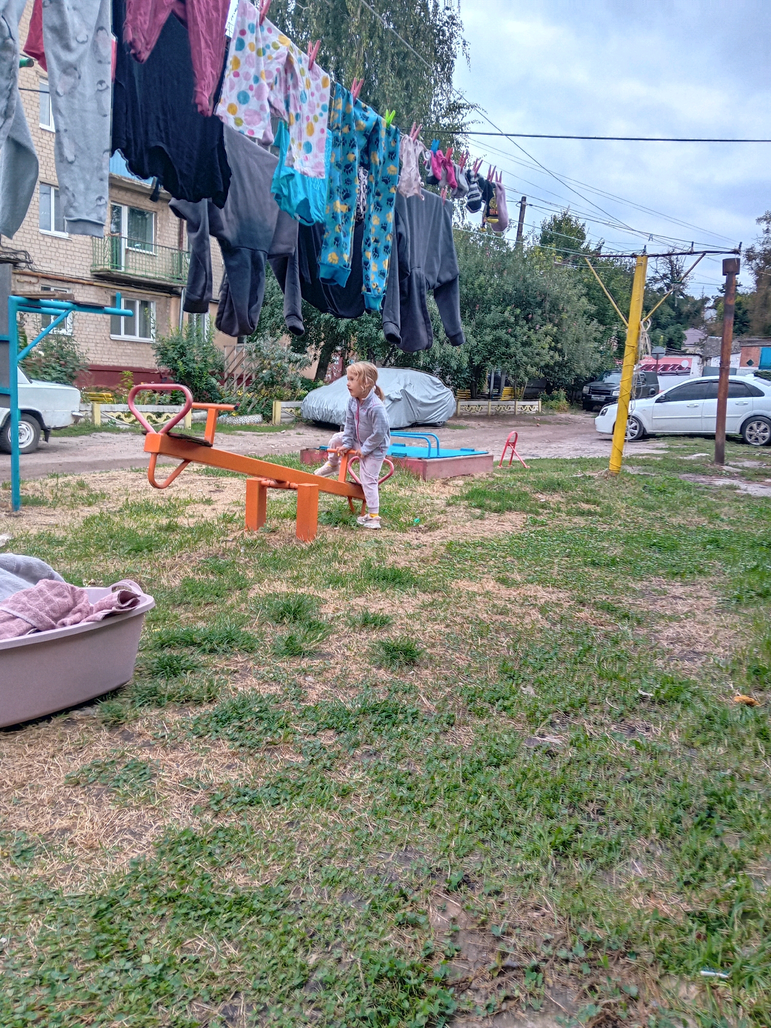 The image captures a scene in the city of Chuhuiv, Ukraine, likely a residential courtyard. In the foreground, a young blonde girl, dressed in light pink sweatpants and a matching zip-up jacket, is seated on an orange seesaw. Her posture suggests she is looking to her right. The seesaw is positioned on a grassy area interspersed with patches of dry earth and clover. To the left of the seesaw, a large, gray plastic tub is visible, containing folded laundry. Clothes hang on a clothesline strung across the frame, some clipped with red clothespins, displaying various colors and patterns, including floral prints and solid dark fabrics. In the background, a two-story brick building with balconies and windows stands to the left, suggesting it is an apartment complex. Beyond the playground area, a sandpit with a blue liner and a small red toy can be observed. Further back, several cars are parked, including a white sedan and a dark SUV. The environment appears to be an outdoor space, with trees and foliage visible throughout. The sky is overcast, contributing to the soft lighting of the scene.