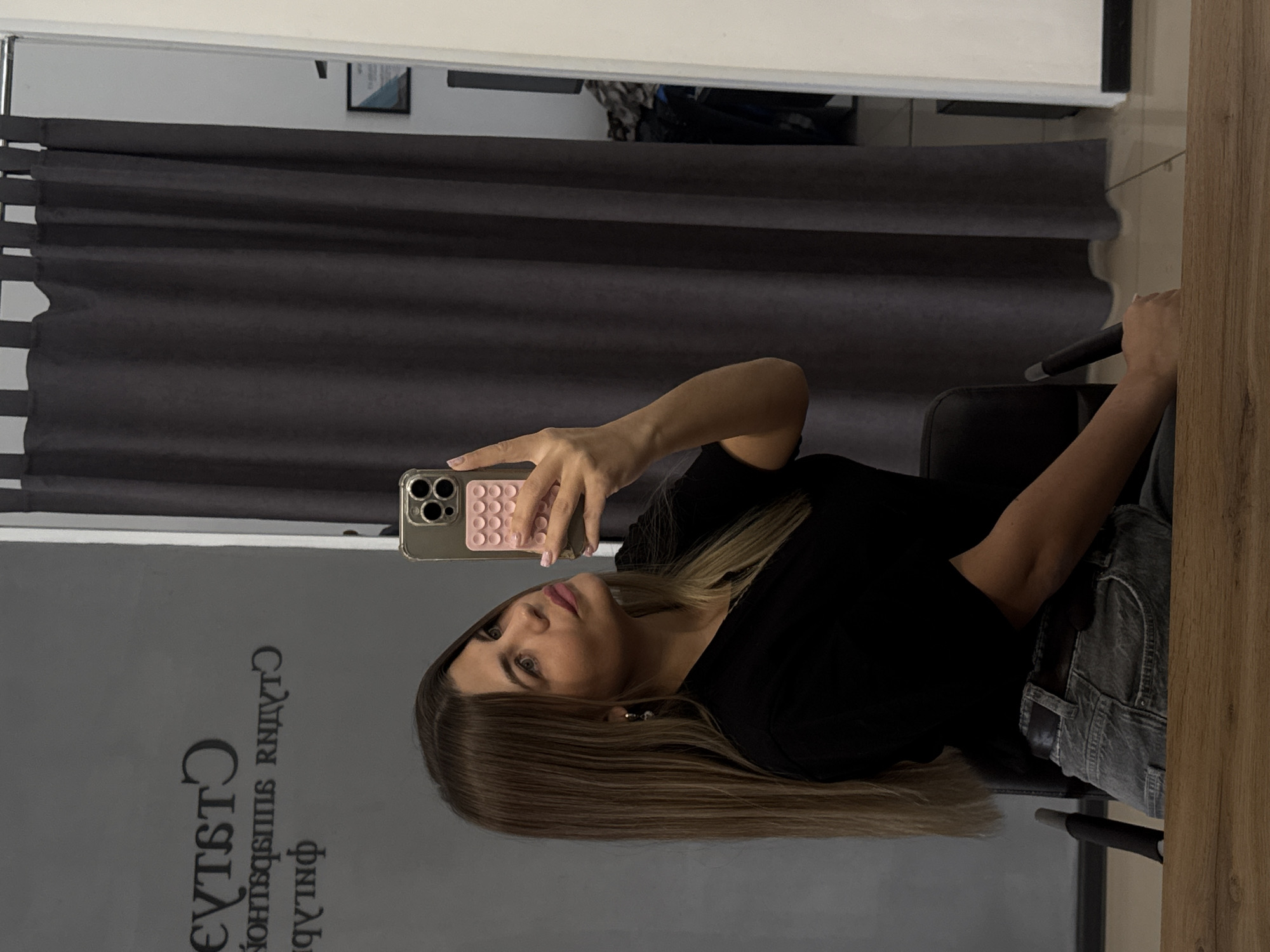 A young woman with long, blonde-brown hair is taking a mirror selfie in a room with a grey curtain and a sign on the wall with Cyrillic writing. She is wearing a black t-shirt and grey jeans. She is holding a smartphone in her right hand and has her left hand in a peace sign. The room appears to be a salon or studio, as indicated by the signage and the presence of a curtain that may be for changing or as a backdrop. The lighting suggests it is indoors during the day. The woman is seated, and the selfie is taken from the chest up, with the background reflecting in the mirror. The sign on the wall reads "Студия" (Studio) in large, dark letters, with smaller text below it. The location is Chaikovsky, Russia.