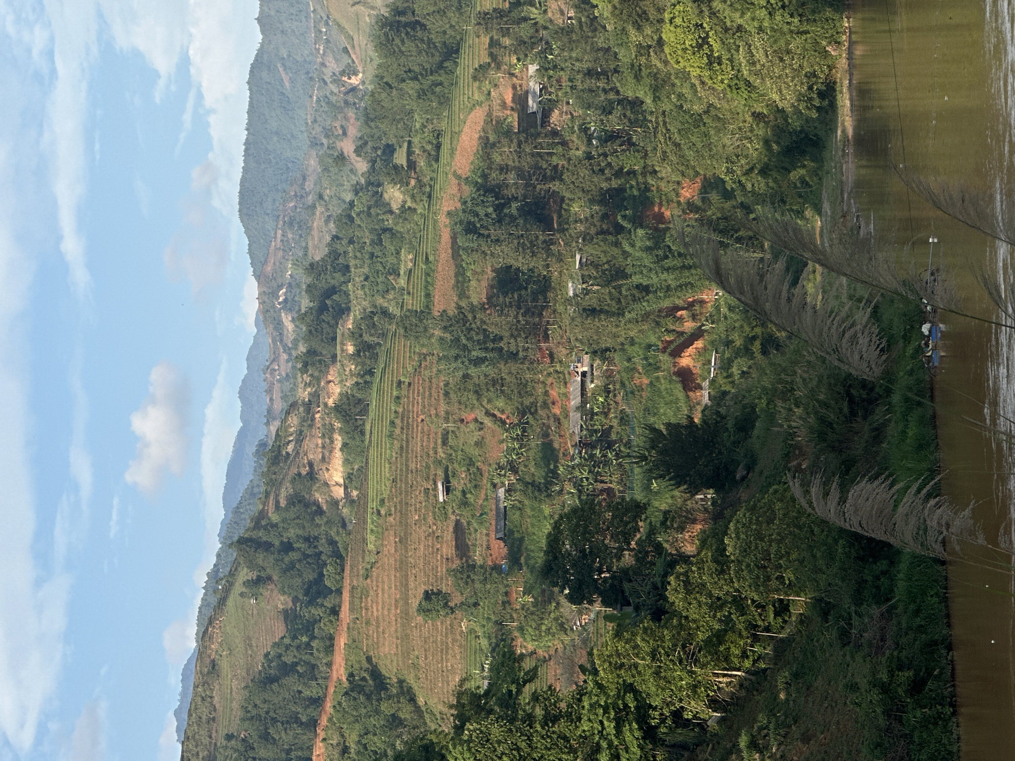 A photograph shows a landscape in the city of Lao Chai, Vietnam. A body of water, possibly a lake or river, occupies the right side of the frame, with vegetation growing along its edge. To the left, a hillside is covered with agricultural terraces. The terraces are interspersed with trees and other greenery. A few buildings or structures can be seen scattered among the terraces. In the background, distant mountains are visible under a sky with scattered clouds.