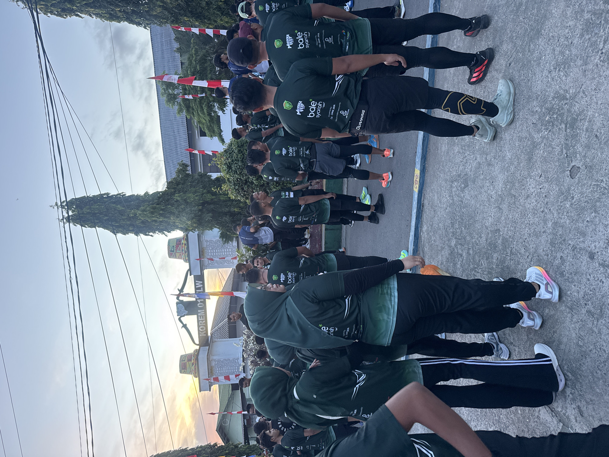 The image depicts a group of people, mostly young men and some women, gathered outdoors in what appears to be a city street in Lhokseumawe, Indonesia. They are dressed in dark green t-shirts with white and light green graphics, suggesting they are part of an organized event, possibly a run or a festival. The t-shirts have text that reads "Merdeka Fest" and "Bale Syariah". Some individuals are wearing headscarves. The backdrop features a building with a prominent archway labeled "KOREM 01/LW", which is likely the Indonesian Army's Military Resort Command 01/Lhokseumawe. Indonesian flags are visible, attached to poles and fluttering in the breeze. The sky is overcast with a warm glow on the horizon, indicating either sunrise or sunset. The ground is paved with concrete. The overall scene suggests a community gathering or event taking place in a public space in Lhokseumawe.