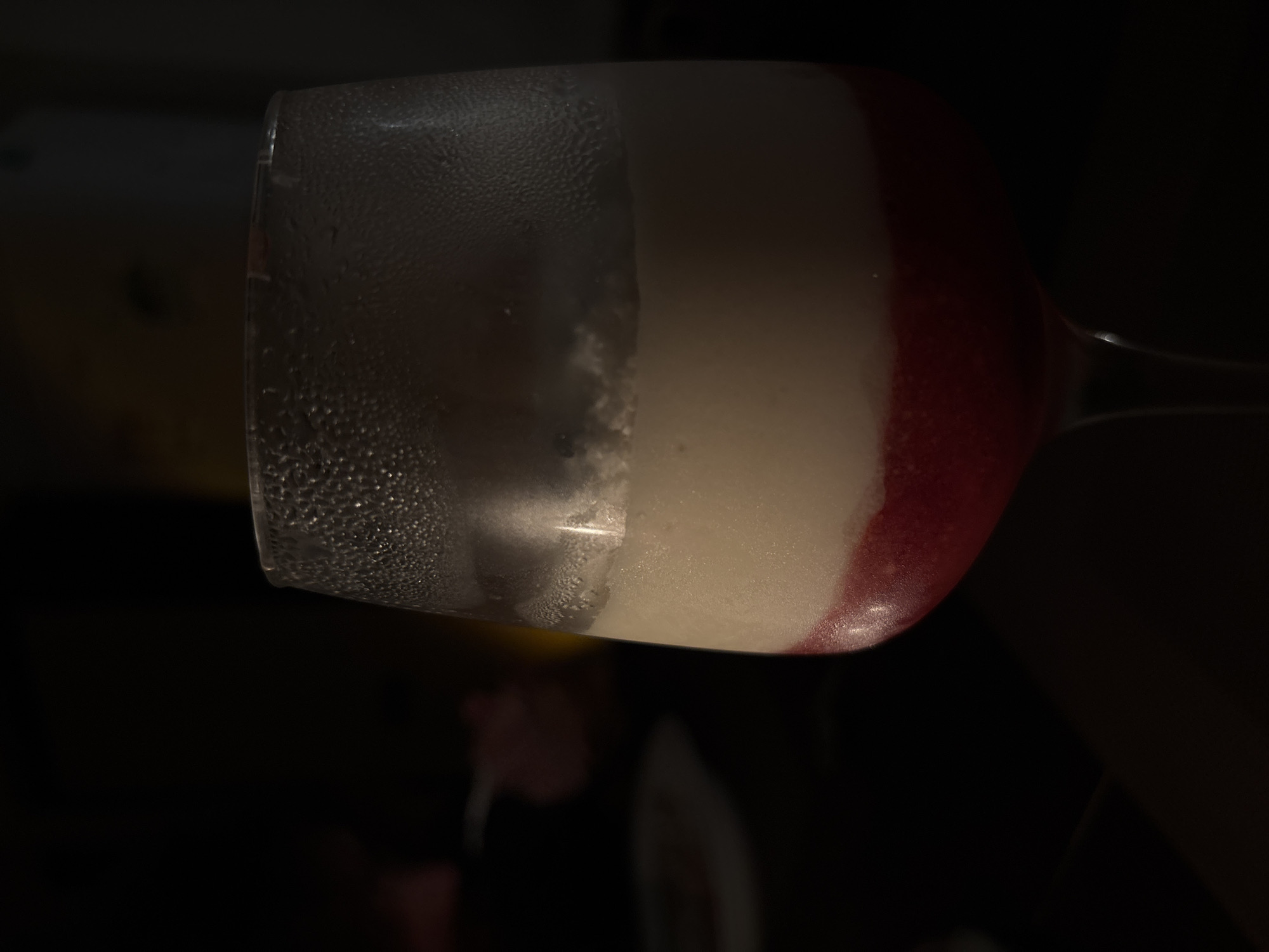 A close-up, low-light, vertical shot focuses on a wine glass containing a layered dessert. The glass is frosted on the exterior, with condensation visible. The dessert has two distinct layers: a bottom layer of vibrant red, likely a fruit puree or jelly, and a top layer of creamy white, appearing to be a mousse or panna cotta. The background is largely dark and out of focus, with hints of color suggesting a dimly lit indoor setting. No people or specific locations in Bečići, Montenegro are discernible.