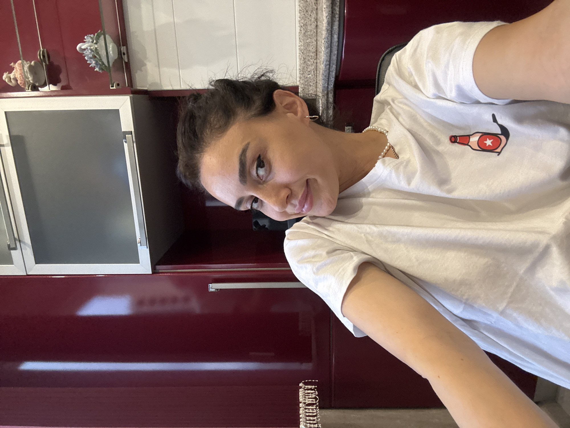 A young woman with dark hair tied back is photographed in a kitchen. She is wearing a white t-shirt with a cartoon image of a beer bottle printed on it and a pearl necklace. The woman is smiling and looking at the camera. The kitchen features dark red cabinets and a grey countertop. A decorative swan figurine and another figurine are visible on a shelf above the countertop. The overall impression is a casual and candid shot taken within a domestic setting.