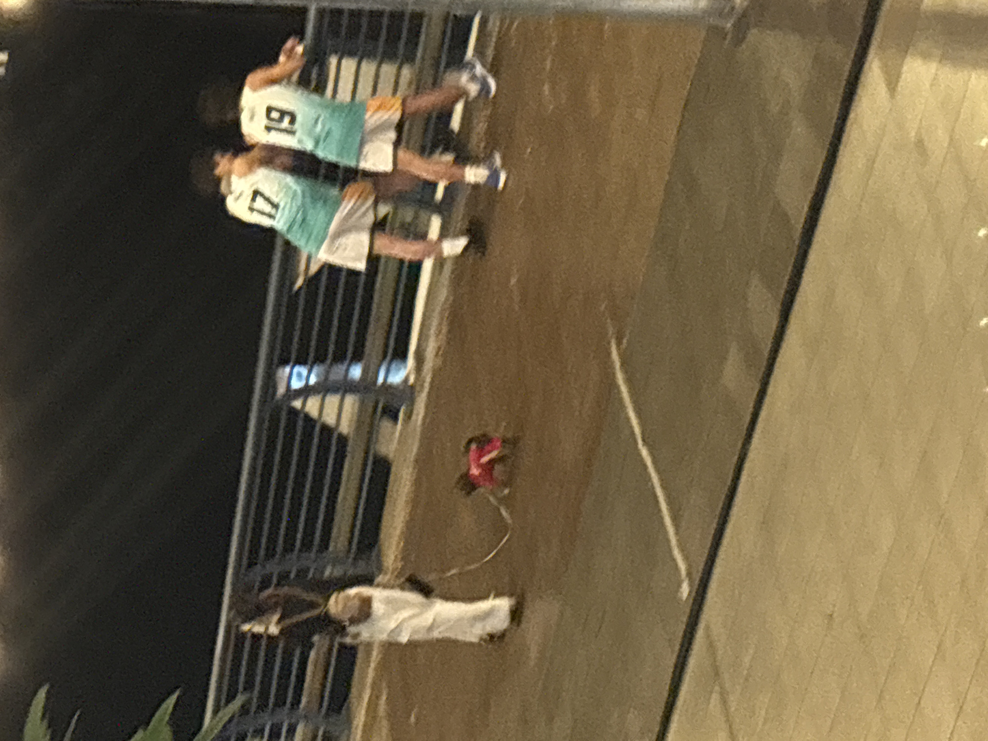 The image captures a scene in North Jakarta, Indonesia. Two individuals wearing matching basketball jerseys numbered 17 and 19 stand near a metal railing. A person dressed in white robes stands several feet away, with a small dog on a leash near them. The ground is paved, and there is a wall or barrier nearby. The lighting suggests it is nighttime.