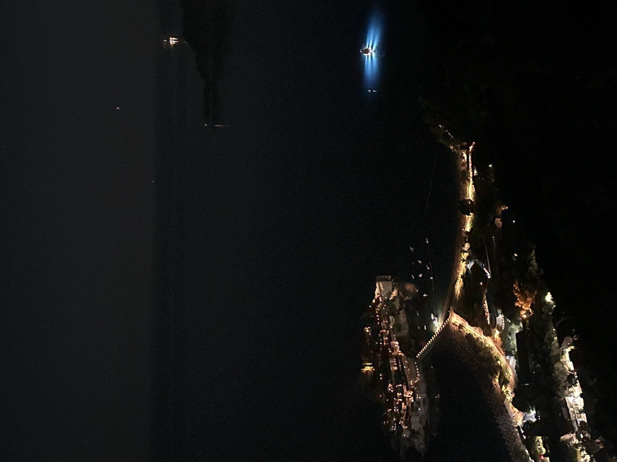 This is an aerial, nighttime photograph of Sveti Stefan, Montenegro. The image is dominated by darkness, with illuminated buildings and lights on a narrow causeway forming the primary visual elements.
In the foreground, on the right side, a dark, mountainous landscape with scattered lights from buildings descends into the foreground. A pathway with numerous small, yellow lights snakes its way down from this elevated viewpoint.
To the left, across the causeway, is the island of Sveti Stefan. The island is densely packed with illuminated buildings, appearing as a cluster of warm, golden lights against the dark sea. The iconic stone buildings are visible, with lights emanating from windows and rooftops. A long, linear string of lights extends from the island along the causeway, connecting it to the mainland.
In the body of water to the right of the island, a single bright blue light illuminates the sea, suggesting a boat or a submerged light source. Further in the distance, faint lights are visible on the horizon, indicating other structures or boats at sea, and the dark silhouette of landmasses. The overall scene is one of a picturesque, illuminated coastal resort at night.
