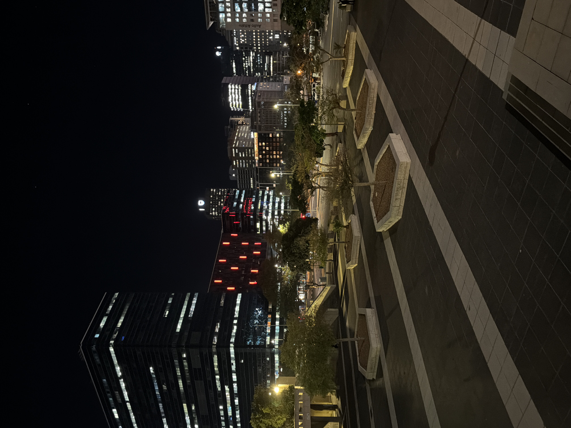 This nighttime photograph captures a view of the city of Cape Town, South Africa. The shot appears to be taken from an elevated vantage point, possibly a pedestrian walkway or balcony. The foreground consists of a paved surface with white striped patterns. Along the left edge of the pavement, there is a row of irregularly shaped planters with small trees.
In the background, the city skyline is visible, illuminated by artificial lights. Tall buildings dominate the scene, with numerous lit windows visible in the skyscrapers. A notable building features a prominent "D" logo on top. Additionally, a building with red lights arranged in a pattern stands out. The overall impression is one of a modern, bustling urban center at night. No people are visible in the frame.