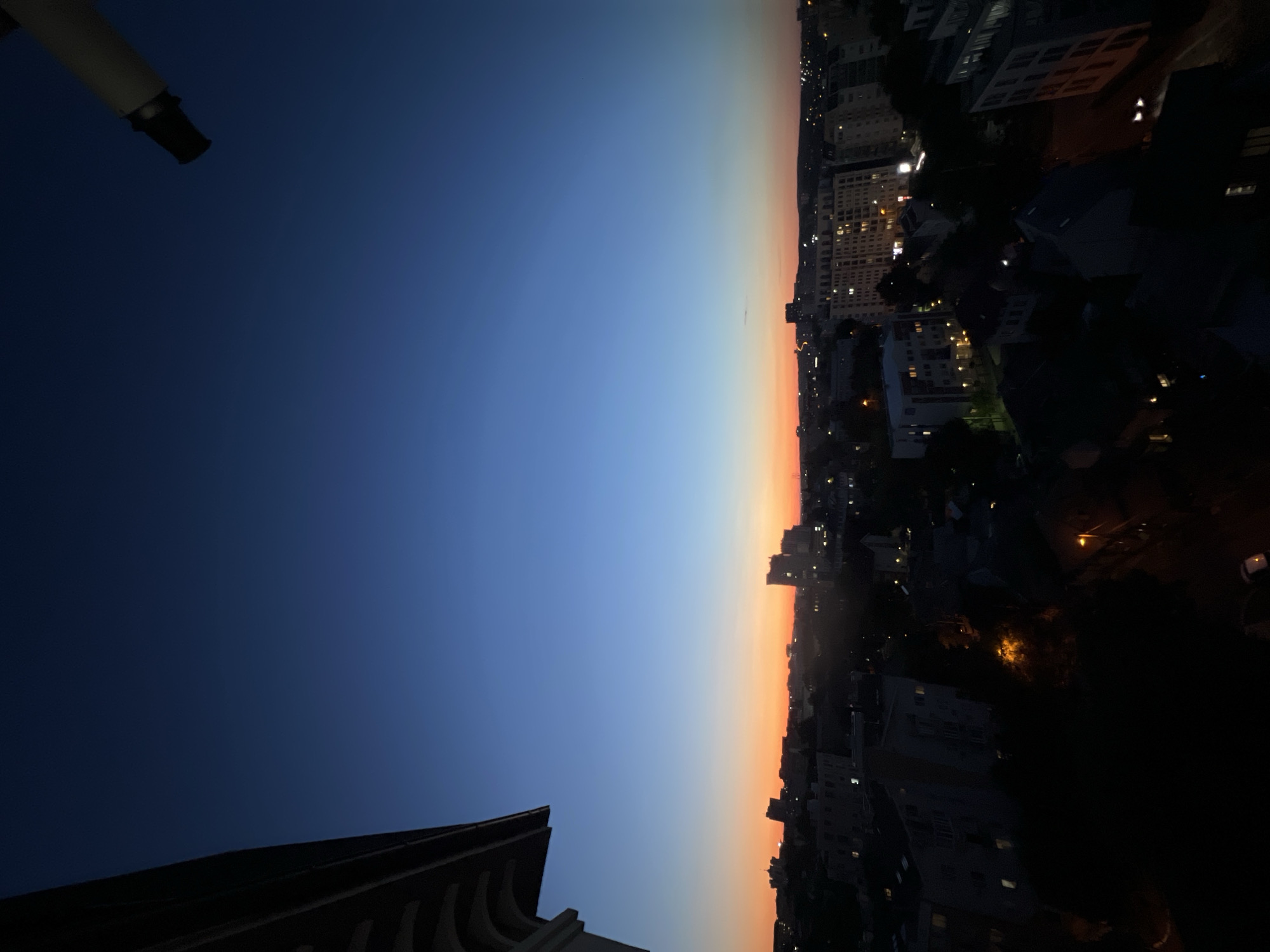 This image captures a twilight view of a city skyline, likely Chișinău, Moldova, given the context. The sky displays a gradient of deep blue at the top, transitioning through lighter blues and then to a vivid orange and pink hue near the horizon, indicative of sunset or sunrise. Silhouetted against this colorful sky are numerous buildings, ranging from smaller residential structures to taller apartment blocks and some imposing office or public buildings. The city lights are beginning to illuminate, with small points of light visible throughout the urban landscape, suggesting the time is early evening or dawn.
In the foreground, on the left side of the image, a portion of a building's architectural detail is visible. This features an ornate, curved overhang or cornice, rendered in dark tones, suggesting it's part of a balcony or roofline. In the upper right corner, a portion of a telescope or similar optical instrument is visible, angled downwards towards the cityscape.
The lower portion of the image is dominated by the dark silhouettes of buildings and trees. The overall impression is one of a peaceful transition from day to night (or vice versa) over a developed urban area. No people are discernible in the image. The scene is static and appears to be taken from an elevated viewpoint.