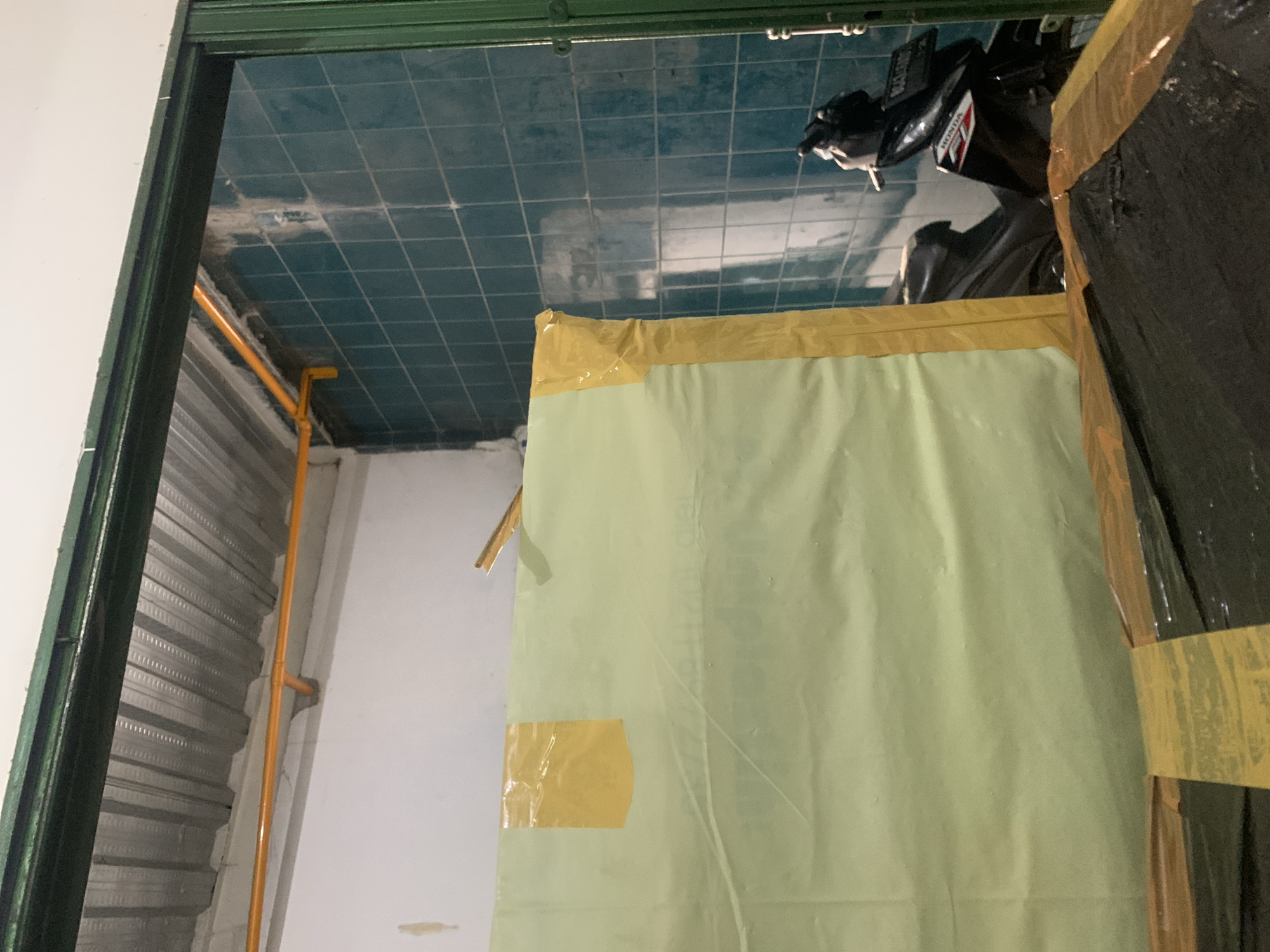The image depicts an interior space, possibly a workshop or storage area, located in Palembang, Indonesia. The scene is dominated by a large, light green tarp secured with yellow tape, which partially obscures the view of the background. To the right, a dark-colored motorcycle is visible, parked against a wall covered in teal-colored square tiles. Above the tiled wall, a white wall and a corrugated metal ceiling are apparent. A bright yellow pipe runs along the wall and ceiling. The overall impression is one of a utilitarian space, possibly undergoing renovation or storage.