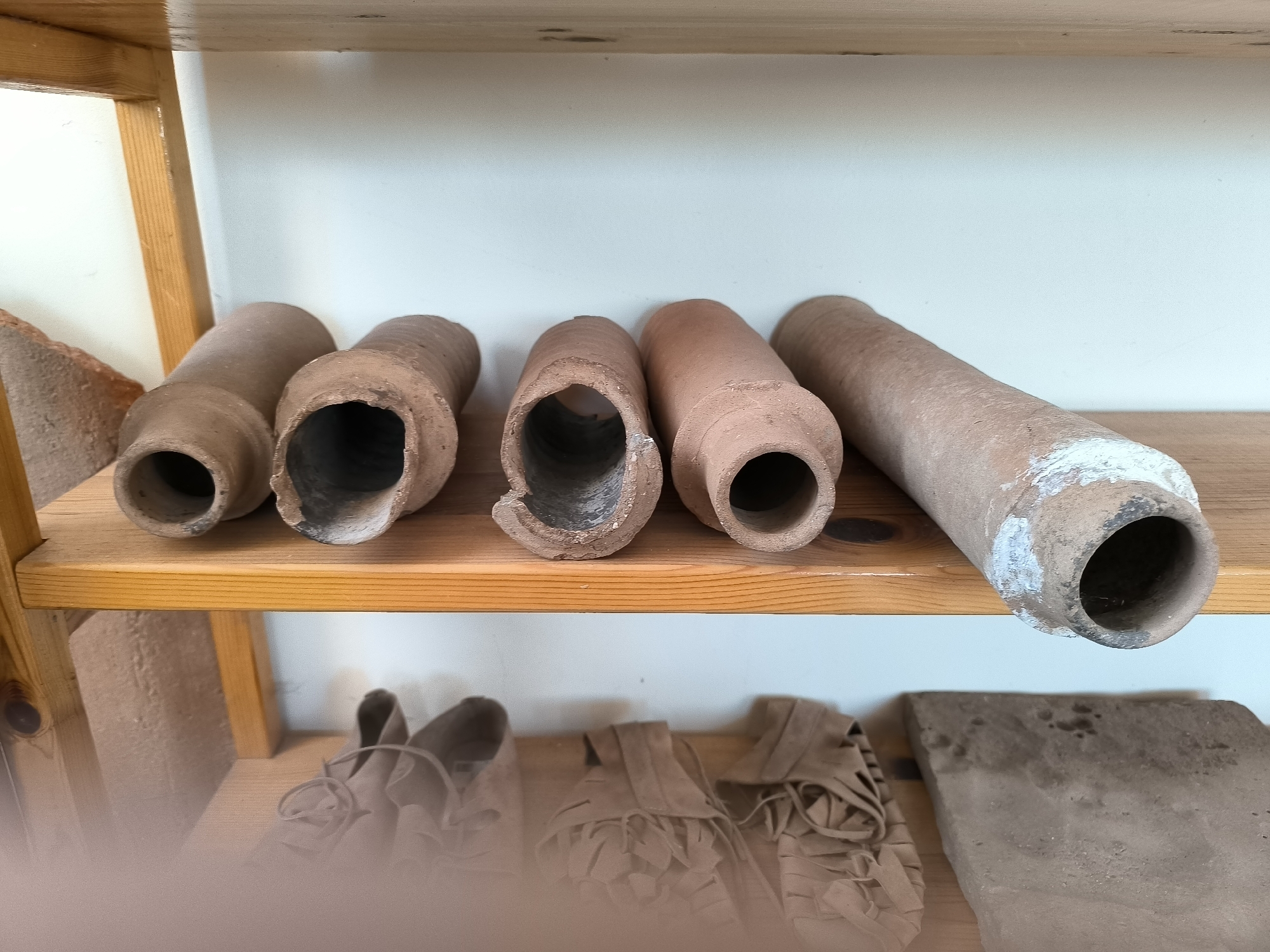 The image shows two wooden shelves against a white wall. The top shelf displays a row of six terracotta pipes of varying lengths and thicknesses. The pipes are positioned horizontally. Some have chipped edges or visible wear. To the left of the pipes, partially visible, is a reddish-brown brick.
The lower shelf contains several items that appear to be made of a light brown material, possibly leather or fabric. These items are arranged from left to right and seem to be footwear or decorative objects with fringe details. To the far right of the lower shelf is a flat, rectangular object, also appearing to be made of terracotta or a similar material.
The scene is likely an indoor setting, possibly a museum, storage area, or exhibition space, given the display of artifacts on shelves. The lighting is even, suggesting artificial light or diffused natural light. The overall impression is one of an organized display of ancient or historical objects. The image's context points to the Garmen village in Bulgaria, known for its archaeological significance.