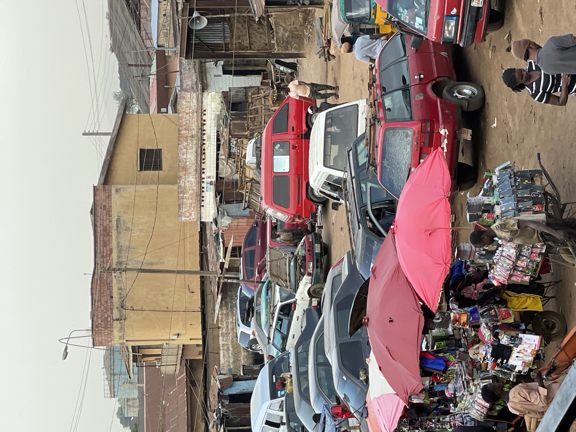 This image depicts a street scene in Bukuru, Nigeria. The view is of a narrow dirt street crowded with vehicles parked haphazardly along both sides. Predominantly red and white vans and smaller cars are visible. Between the vehicles and along the edge of the frame are several vendors displaying goods for sale; one is selling accessories from a pushcart shaded by a pink umbrella. Individuals are present, either standing and interacting or tending to their businesses. Buildings, some with dilapidated exteriors, line the street, with overhead wires crisscrossing the scene. The sky is overcast.