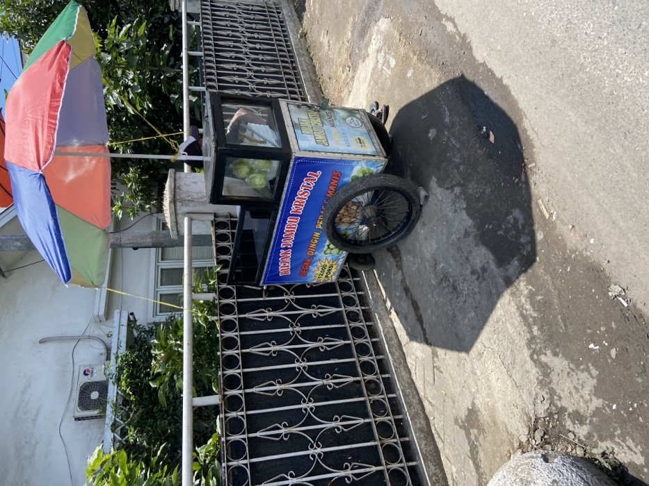 A street vendor is selling "Minuman Jambu Kristal" (crystal guava drink) on the side of a street in Padalarang, Indonesia. The vendor is sitting under a colorful umbrella and has several guavas in a clear plastic container inside the cart. The cart has a blue and white sign with the name of the drink and the price of 12,000 rupiah per portion. There is a white house on the left side of the image, and the street is paved with asphalt. There is a metal fence separating the house from the street. The vendor is wearing a white shirt and black pants. The coordinates provided (latitude: -6.855, longitude: 107.498) are in the city of Padalarang, Indonesia. The shadow of the umbrella is on the street in front of the cart.