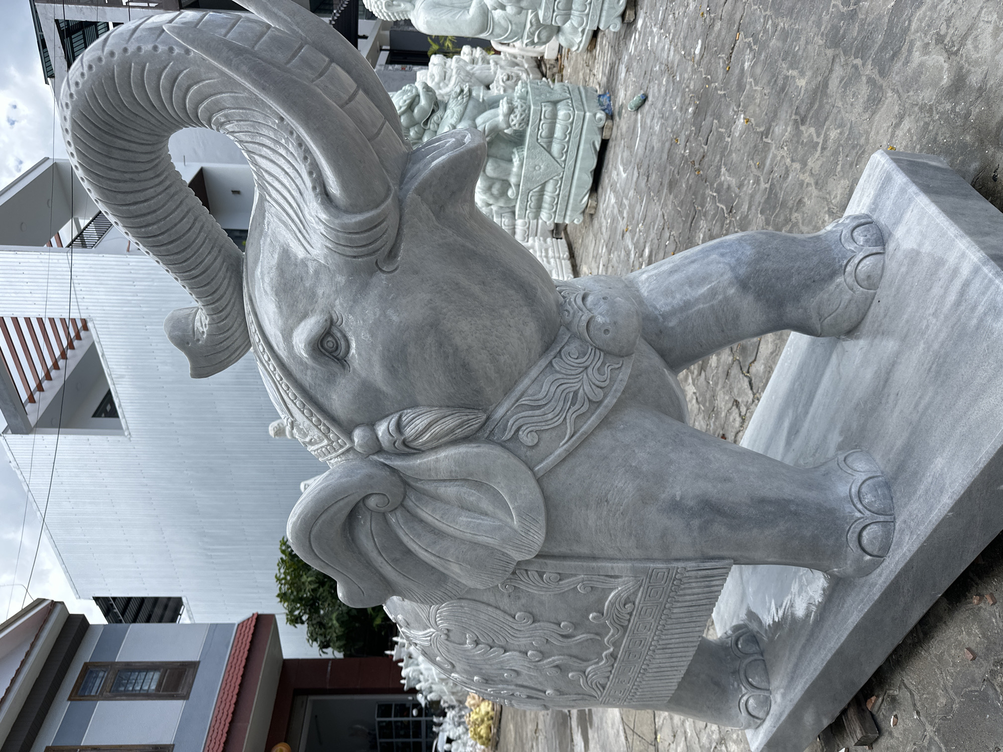 This image captures a large, intricately carved marble elephant statue positioned outdoors in what appears to be a sculpture yard or workshop. The elephant is grey and has detailed carvings on its back, resembling a decorative blanket, and its trunk is raised high, curled upwards. The statue stands on a marble base, which is itself on a paved outdoor surface. In the background, other stone statues are visible, including what appears to be a Buddha statue and some guardian lion figures. Buildings with modern architectural styles, including corrugated metal siding and balconies, are present in the background, suggesting an urban or semi-urban setting. The sky is overcast, indicating a cloudy day. The location is identified as Đà Nẵng, Vietnam.