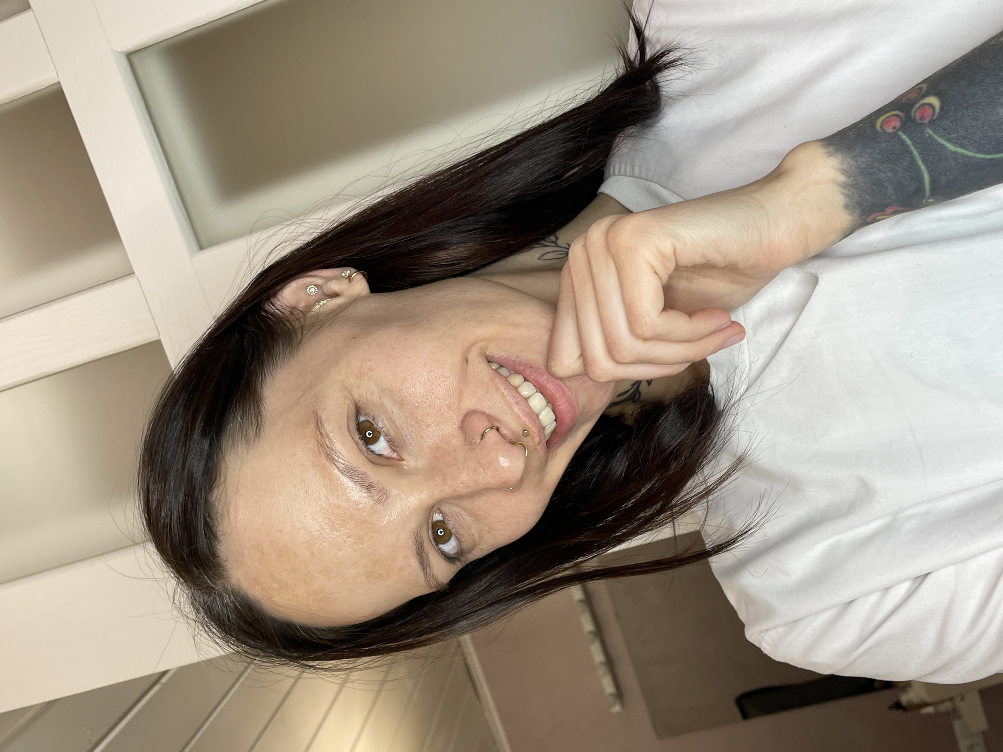 A woman with dark brown hair and multiple piercings in her ear and nose is smiling at the camera. She has tattoos on her left arm and neck. She is wearing a white t-shirt. The background includes a white door or wall panel. The image is oriented in portrait format. It is unlikely that there are details in the image to place the subject in Izhevsk, Russia.