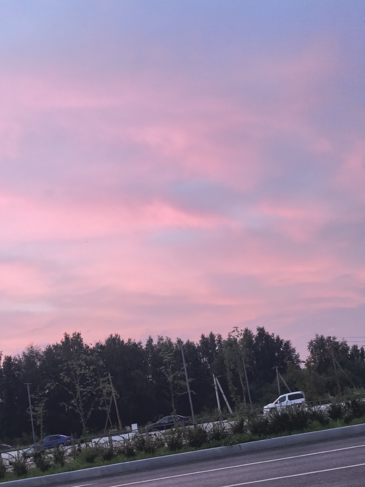 The image shows a dusky sky with shades of pink and purple, likely due to sunset or sunrise. A line of dark green trees forms a horizon against the colorful sky. In the foreground, a paved road with white lane markings is visible. Several vehicles, including a blue car and a white van, are on a road running parallel to the trees. The scene appears to be set in a semi-urban or suburban area, possibly near a highway or main road. The overall atmosphere is serene and natural.