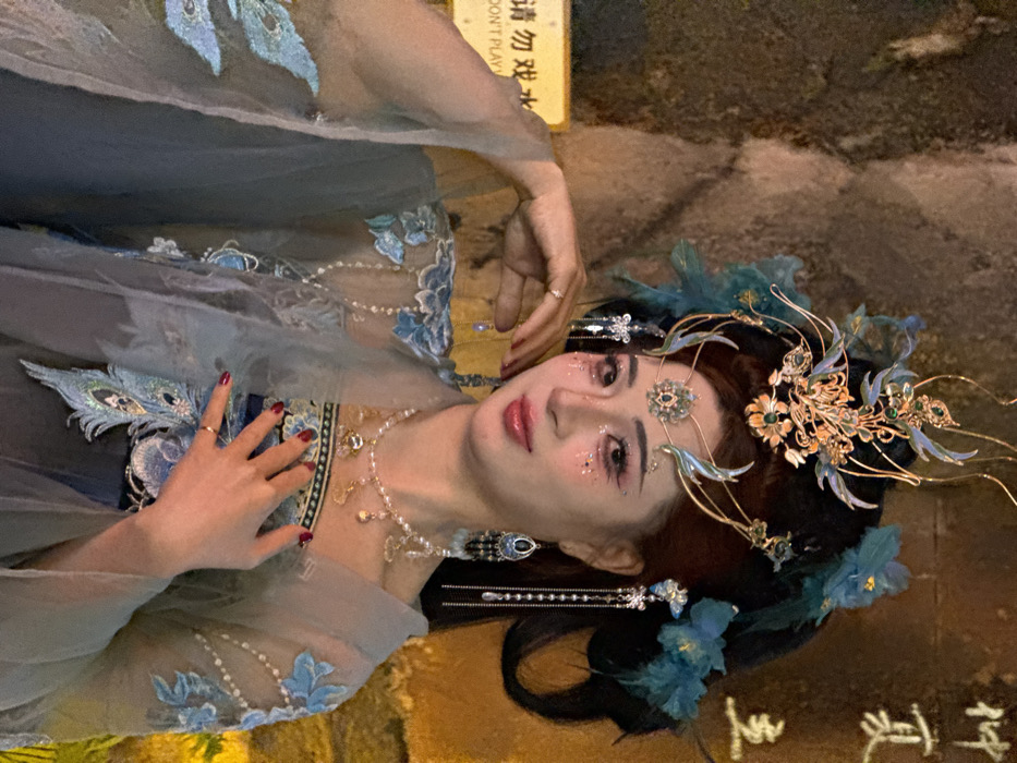 This image depicts an individual posing outdoors at night in Yuzhong District, China. The subject is an adult female with fair skin and dark hair styled into an elaborate updo, heavily adorned with a prominent gold and teal headpiece featuring intricate metalwork, gemstones, and feather-like elements. Smaller decorative pieces, including a jeweled forehead band and dangling ornaments, complement the main headpiece. Her face is heavily made up with pink eyeshadow, glitter on her cheeks, and glossy pink lips. She wears long, multi-tiered earrings, one appearing dark blue/black with beads and the other silver with chains, along with a pearl necklace featuring multiple small golden charms. Her fingernails are painted red, and she wears a simple ring on her left ring finger.
The woman is dressed in traditional-style attire, consisting of layered, sheer light blue or grey-blue fabric with intricate embroidery. Visible embroidery includes blue floral motifs on the sleeves and a prominent peacock feather design on the torso. Her left hand is raised with fingers near her temple, while her right hand rests on her chest, near the peacock embroidery. She gazes directly forward with a slight smile.
The background is dark and indistinct, suggesting a night setting, with rough, textured surfaces possibly indicating stone or rock walls. Portions of the background are dimly illuminated by out-of-focus golden light sources. A yellow rectangular sign is partially visible in the lower left, displaying Chinese characters and the English phrase "DON'T PLAY." Additional faint Chinese characters are discernable on the upper right background.