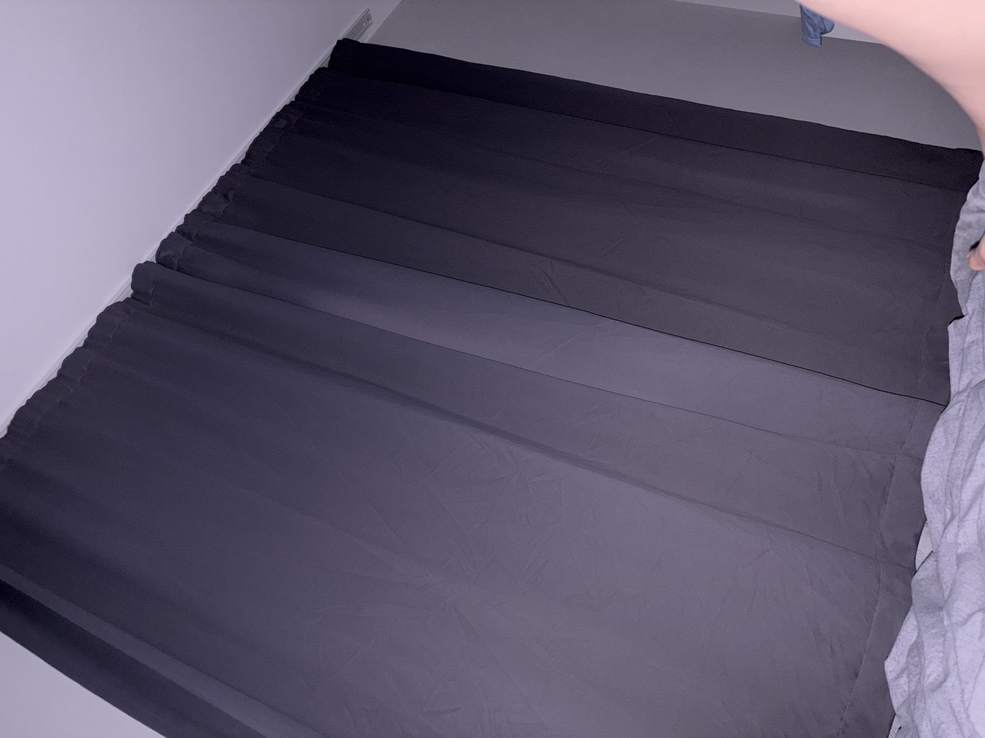The image shows a close-up of dark gray curtains hanging from a track against a white ceiling. Below the curtains, a white, textured surface, likely a bedspread or duvet, is visible. The lighting appears dim and possibly artificial, casting subtle shadows on the fabric of the curtains. The scene is indoor and domestic, with no discernible elements that would indicate a specific location within Stara Zagora, Bulgaria. The focus is entirely on the textures and folds of the fabric and the ceiling.
