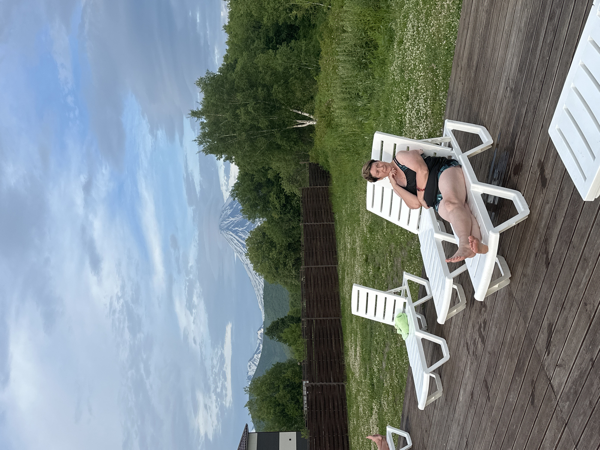 A woman is lying on a white plastic chaise lounge on a wooden deck. She is wearing a black swimsuit with a patterned trim and has her head resting on her hand, with her eyes closed. The deck is surrounded by lush green grass with scattered white wildflowers. In the background, a wooden fence separates the grassy area from a dense forest of trees. Beyond the trees, snow-capped mountains rise under a cloudy sky with patches of blue. The scene appears to be set during the daytime, with the weather looking overcast. The overall impression is one of relaxation and tranquility in a natural setting.