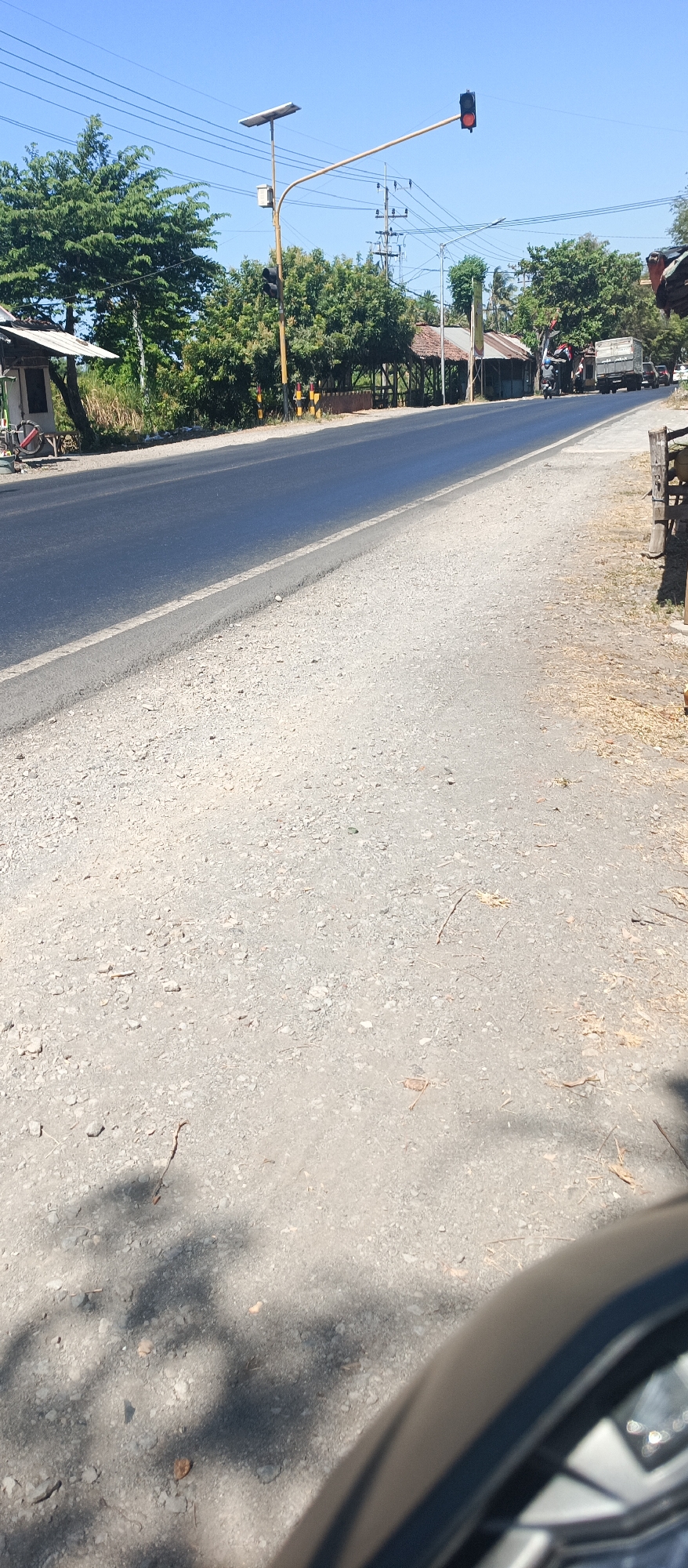 The image shows a rural road scene in Arjasa, Indonesia, under a clear blue sky. A traffic light is visible, displaying a red light. On the left side of the road, there's a small structure with a white roof and some vegetation. The road surface is asphalt, with a gravel shoulder. In the background, trees and some buildings line the road. A few vehicles, including a truck and motorcycles, are present on the road. The lower part of the image shows the shadow of a vehicle, possibly a car, with a portion of its tire and wheel visible.