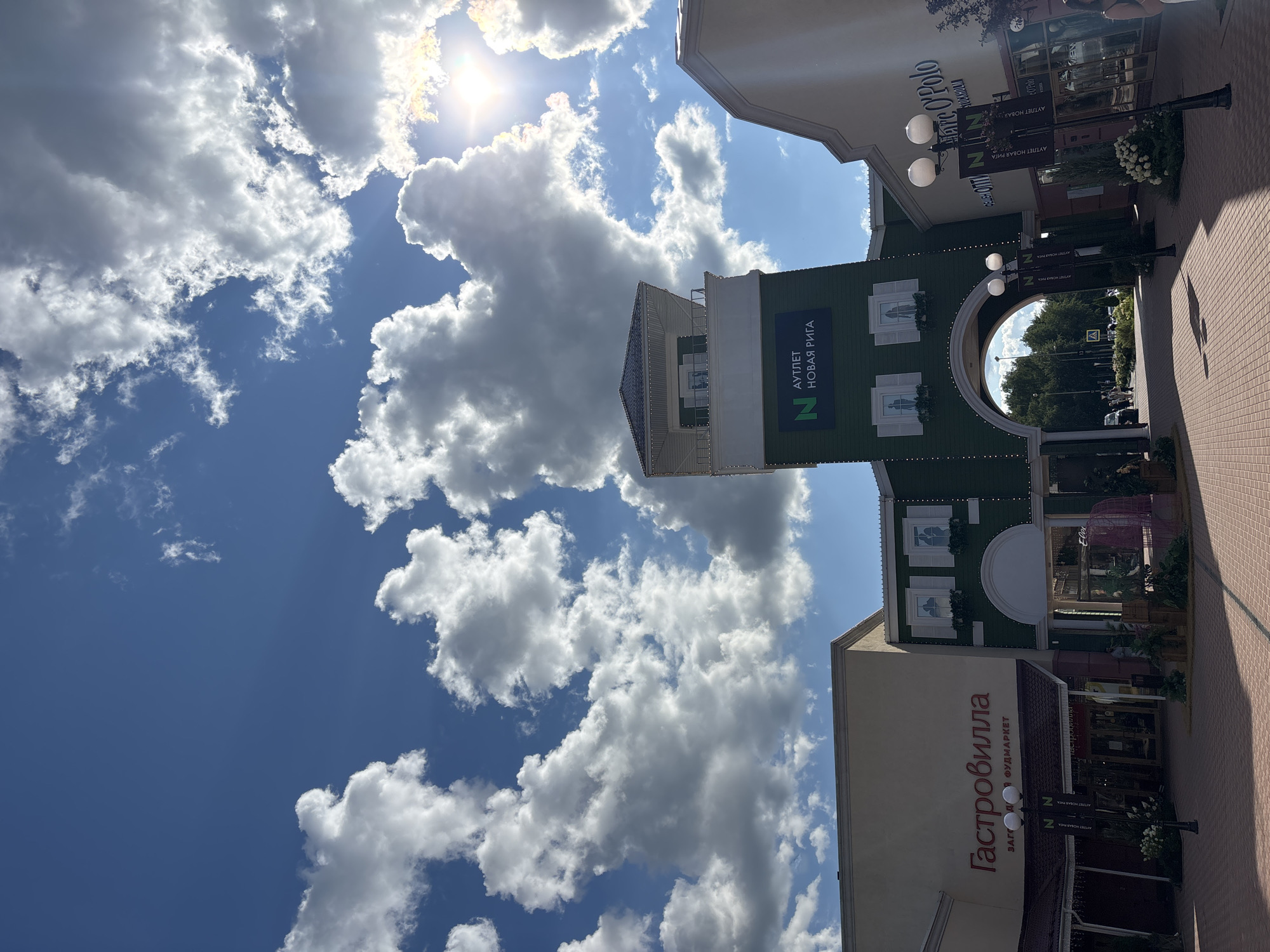 The image shows an outdoor scene at the Novaya Riga Outlet in Zakharovo, Russia. The sky is blue with scattered white clouds and the sun is shining brightly. In the foreground, there is a paved walkway leading through an archway between buildings. To the left is a building with the Cyrillic text "Гастровилла" and "ЗАГОРОДНЫЙ ФУДМАРКЕТ" on its facade. To the right, a building displays the "Marc O'Polo" logo. A green tower with a white balcony and the Cyrillic text "АУТЛЕТ НОВАЯ РИГА" (Outlet Novaya Riga) is visible in the center. Several lampposts are positioned along the walkway. In the background, trees and more buildings can be seen. There are a few people walking on the walkway and near the entrances of the shops.