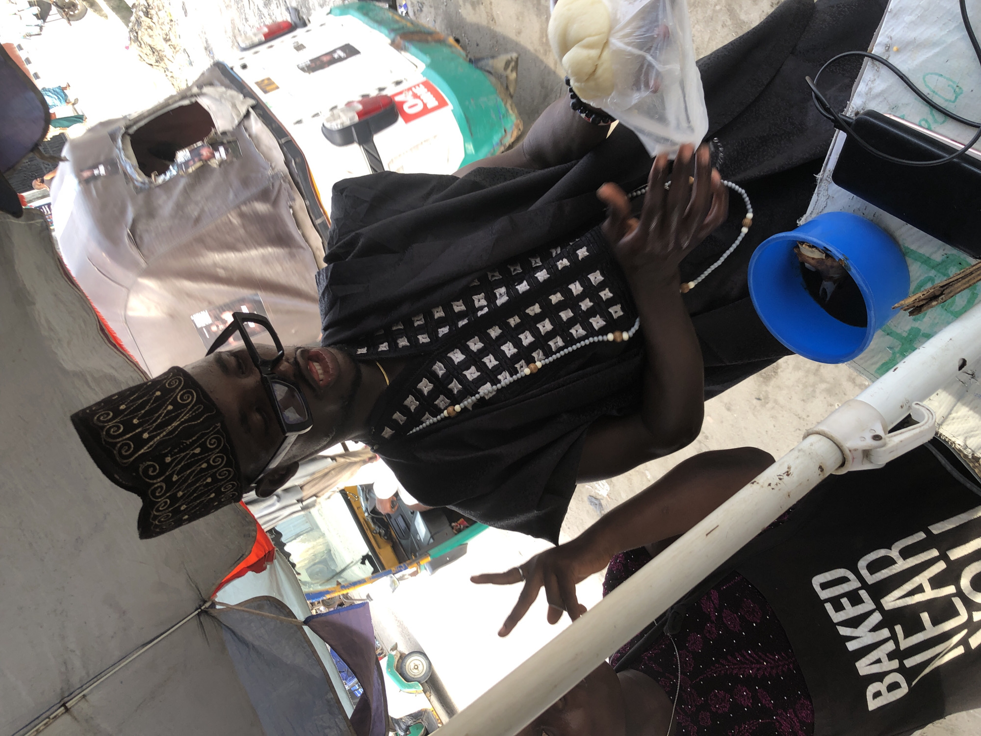 A man wearing a patterned black cap and large glasses is the focal point. He's wearing a black shirt with a white diamond pattern and a necklace of white beads. In one hand, he holds a clear plastic bag containing a white ball. A blue plastic cup containing a dark liquid sits on a surface beside him, along with a black electronic device and charging cable. A white umbrella pole extends into the foreground, partially obscuring a woman wearing a black shirt printed with the phrase "BAKED NEAR". The background shows a busy city street in Ado, Nigeria, with various vehicles, including a white and green bus.
